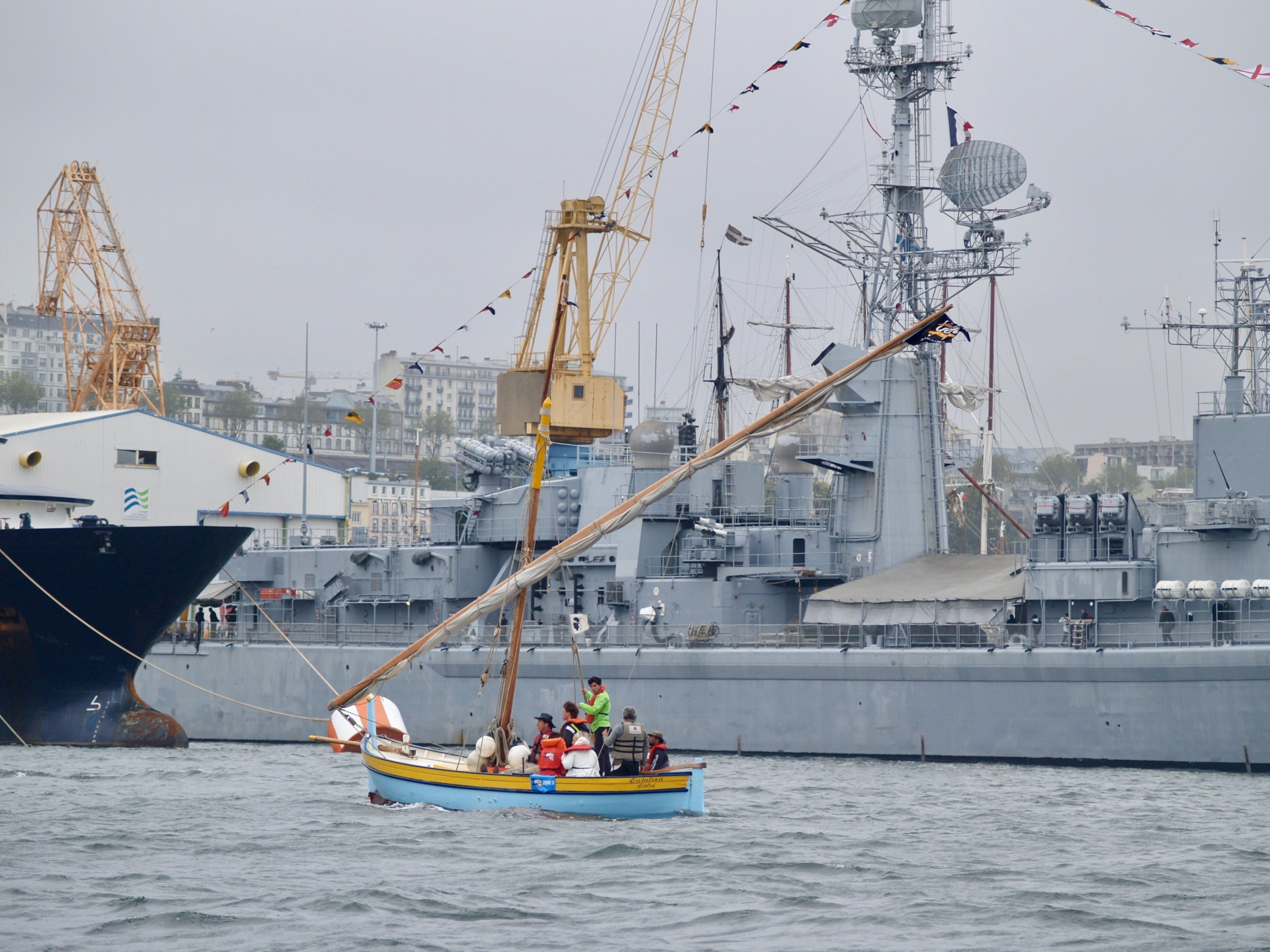 Un petit bateau de plaisance avec plusieurs personnes à bord, dont certains portent des gilets de sauvetage, navigue dans un port. En arrière-plan, on voit un grand navire de guerre ou un porte-avions amarré, avec des structures métalliques, des ante