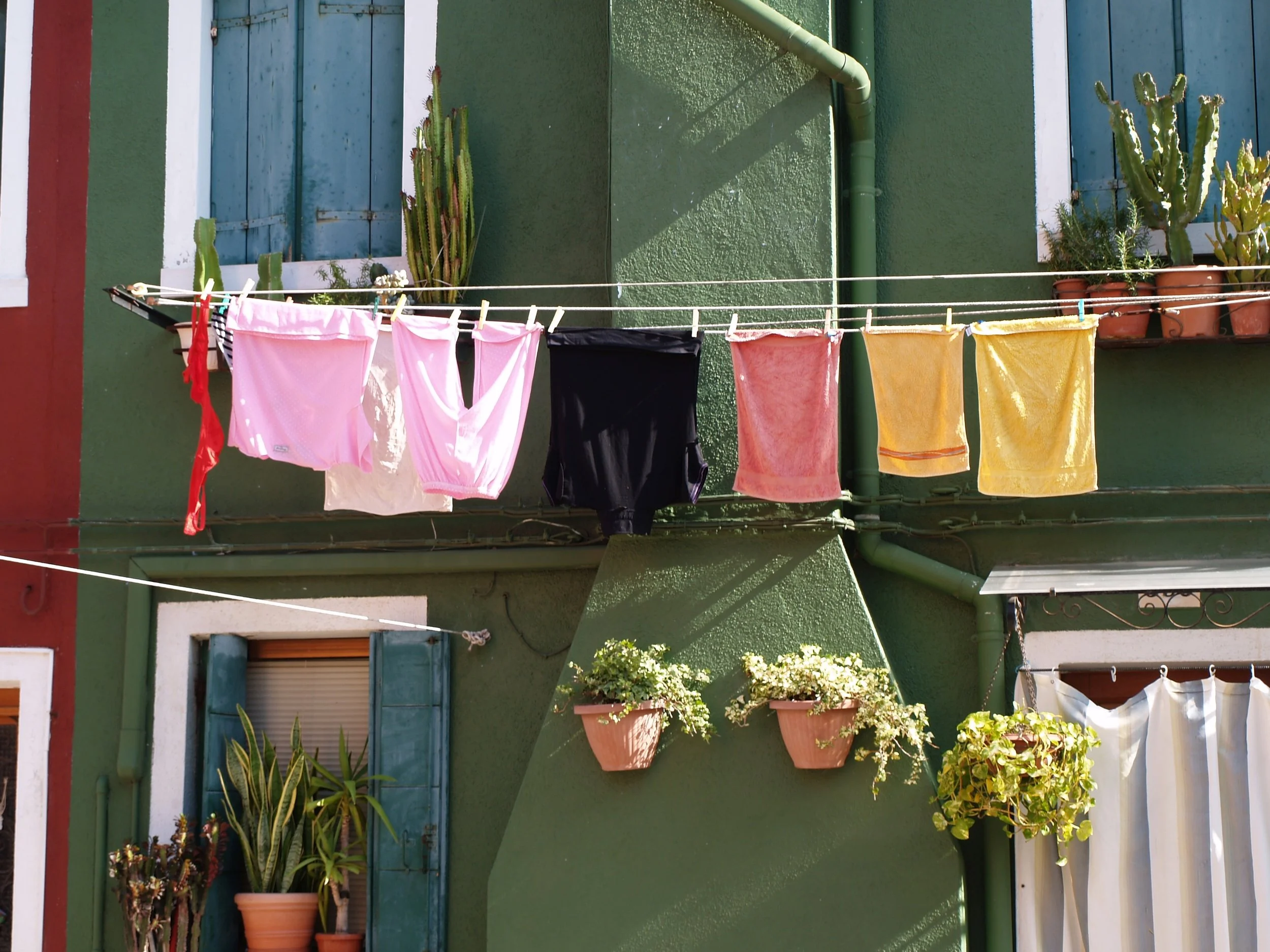 Balcon avec des plantes en pot et des vêtements suspendus en train de sécher. Murs verts, fenêtres avec volets bleus et clôture en métal.
