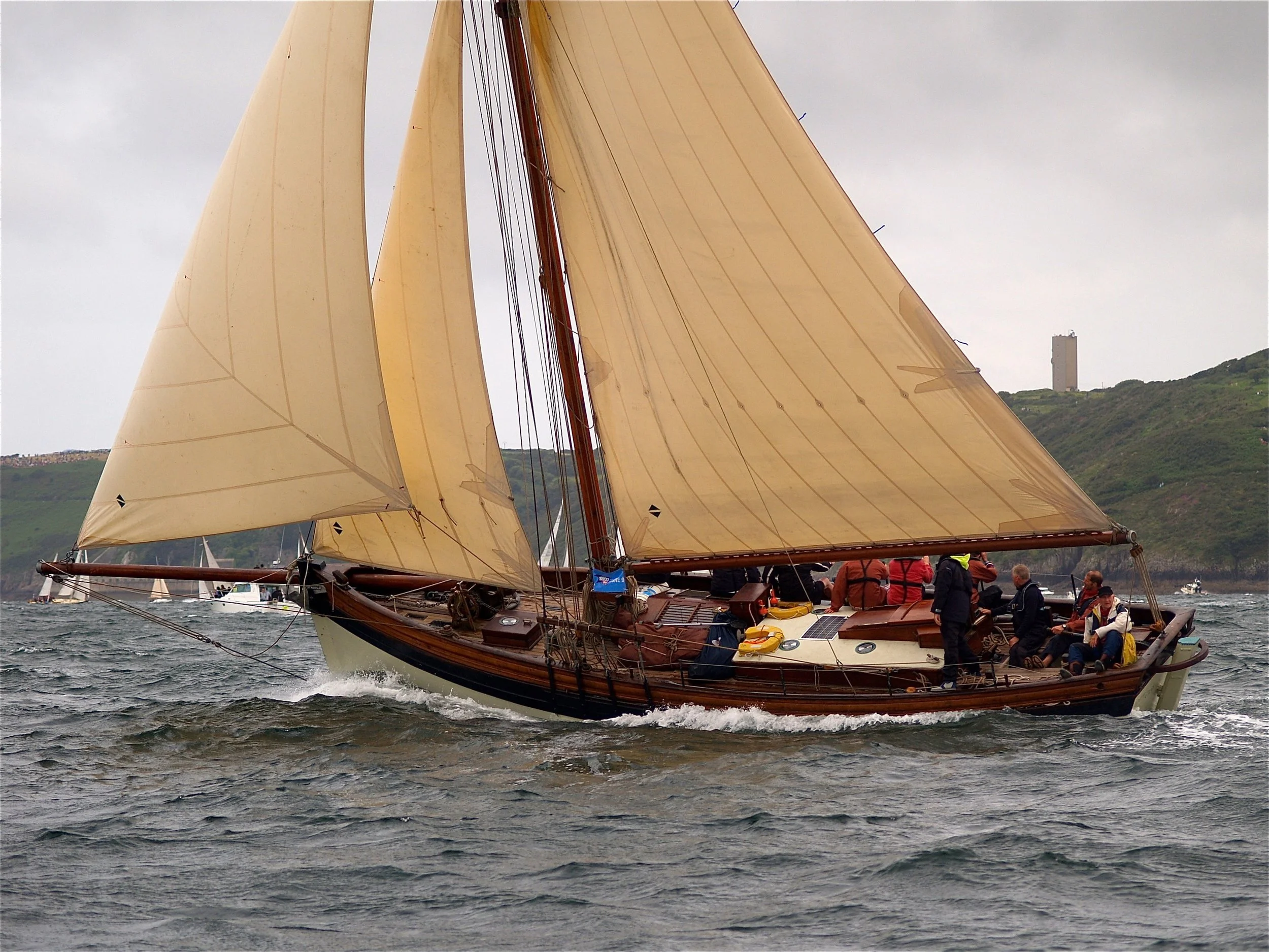 Un voilier en bois avec des voiles en toile beige naviguant sur l'eau, avec des personnes à bord, le paysage en arrière-plan inclut une colline verte et une tour en pierre.