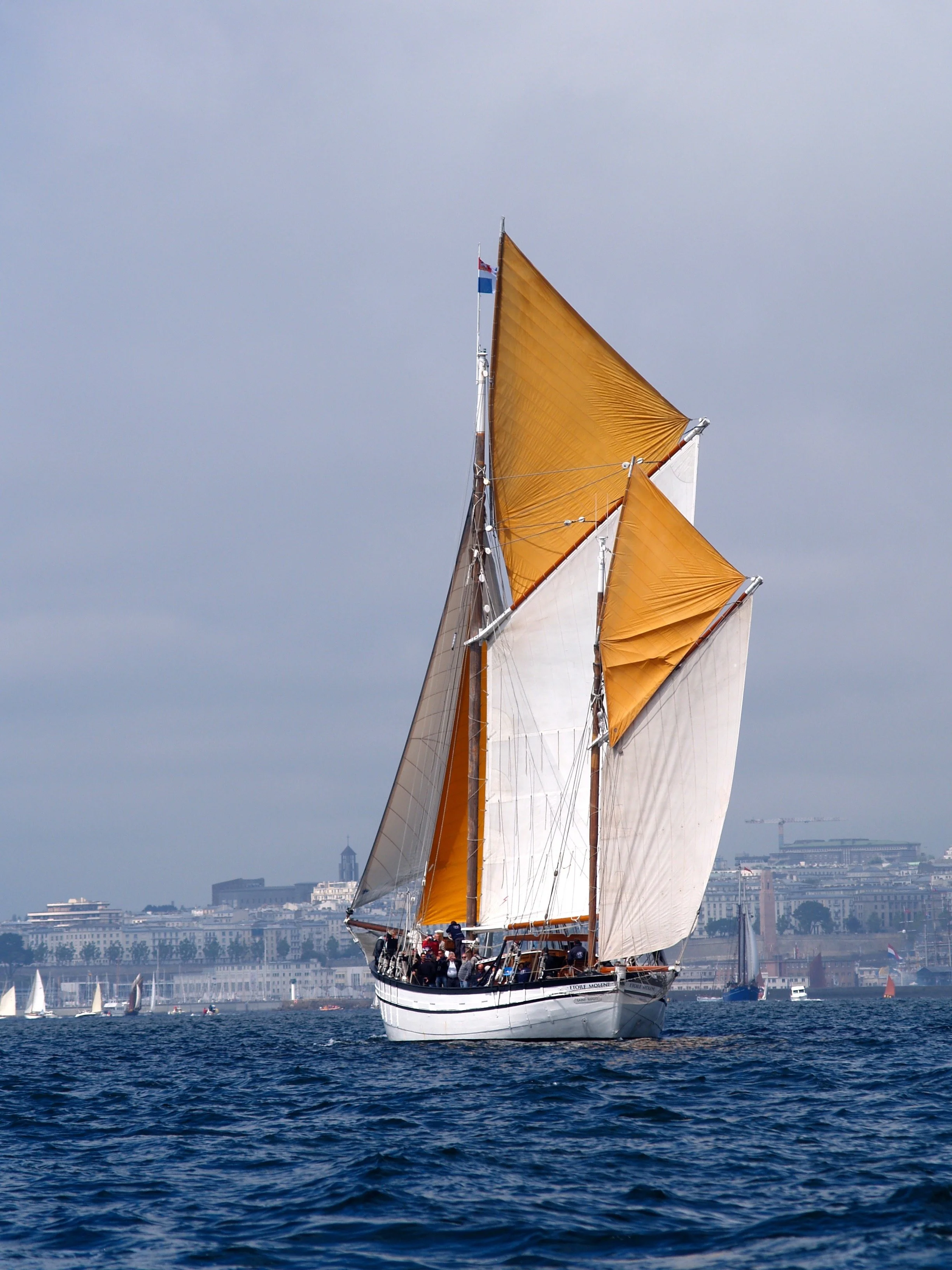 Un voilier avec des voiles orange et blanches navigue sur un fleuve ou une mer, avec une ville en arrière-plan et plusieurs autres petits voiliers autour.