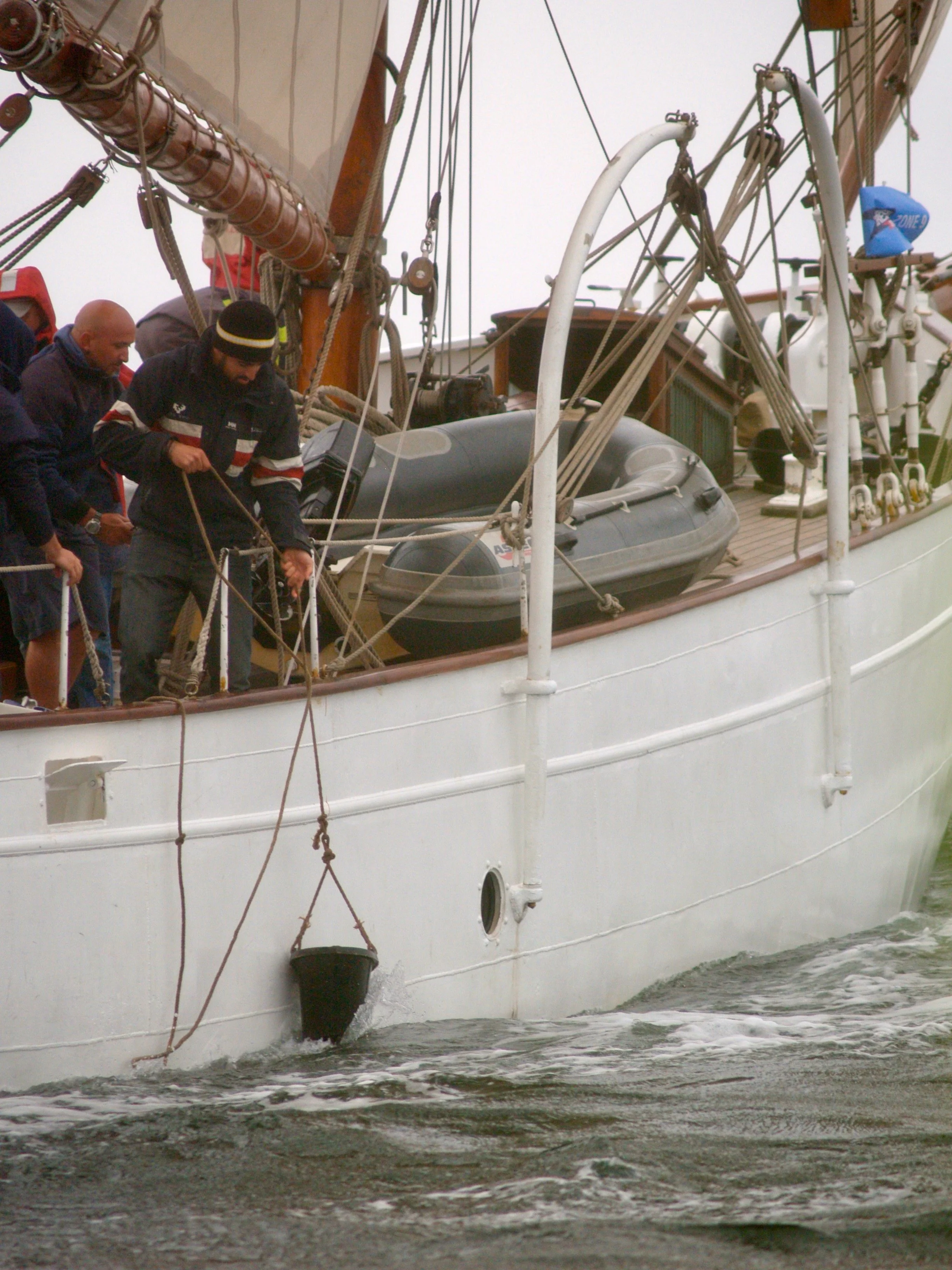 Deux hommes préparent un voilier à quai, un d'eux tient une corde et travaille à l'arrière du bateau, il y a un canot pneumatique noir attaché au côté du voilier.