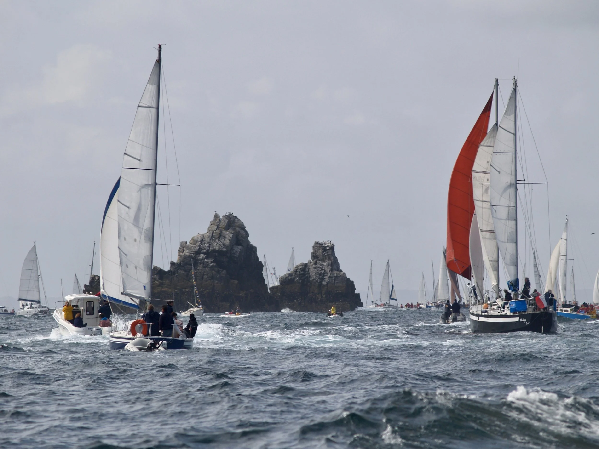 Des voiliers naviguant sur la mer avec un rocher en arrière-plan, des gens à bord et sur la jetée.