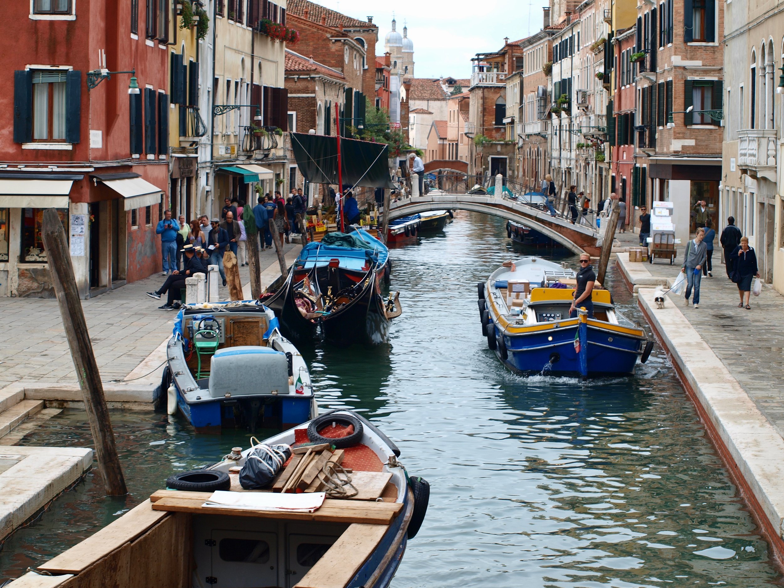Une rue piétonne bordée de bâtiments colorés avec des fenêtres à volets, où des gens se promènent. Au centre, un canal avec des bateaux amarrés et un petit pont enjambant l'eau.