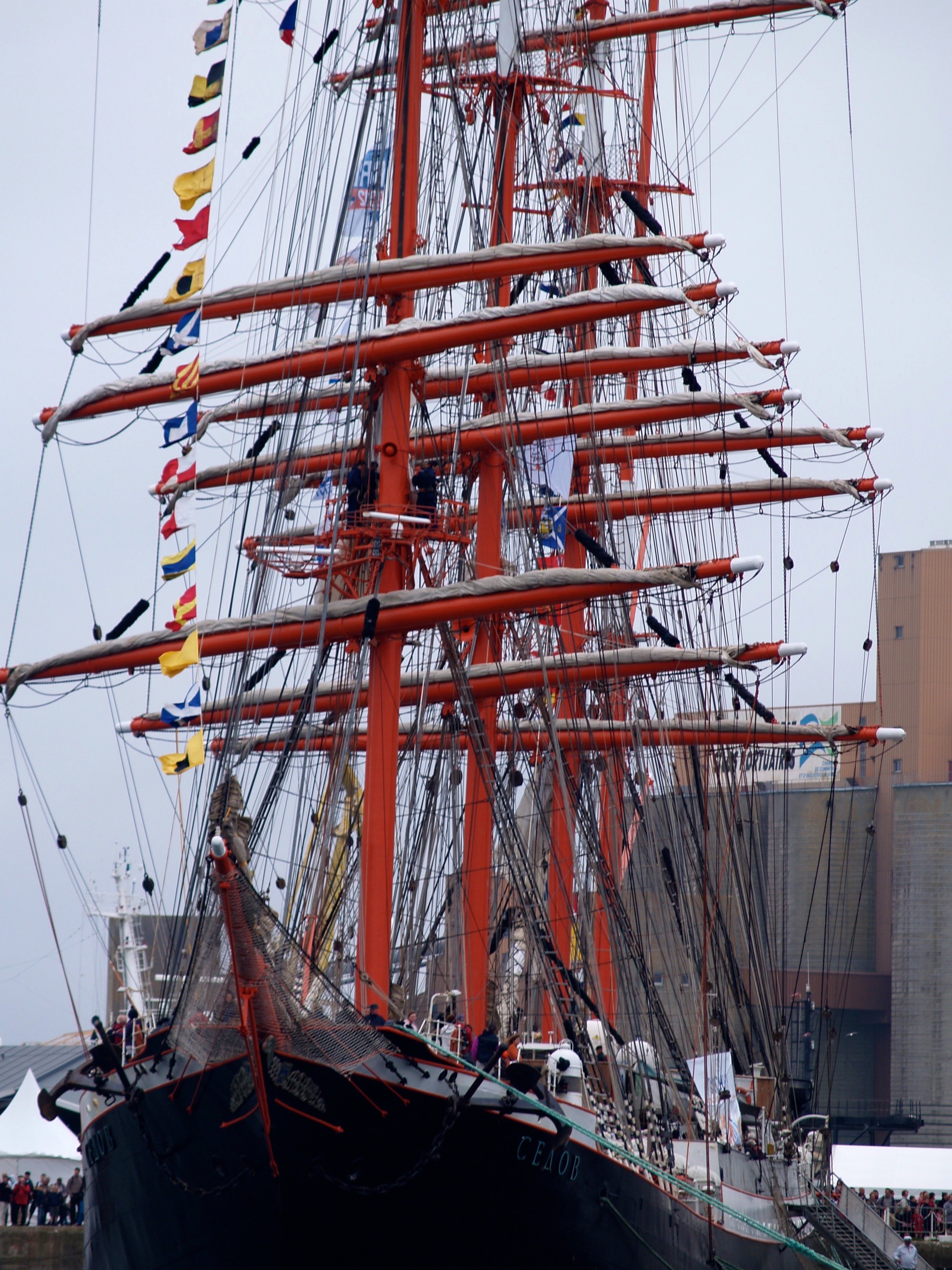 Un grand navire à voiles de couleur noire avec des mâts orange, des cordages, et des drapeaux multicolores, amarré dans un port, avec des personnes devant le bateau et des bâtiments en arrière-plan.