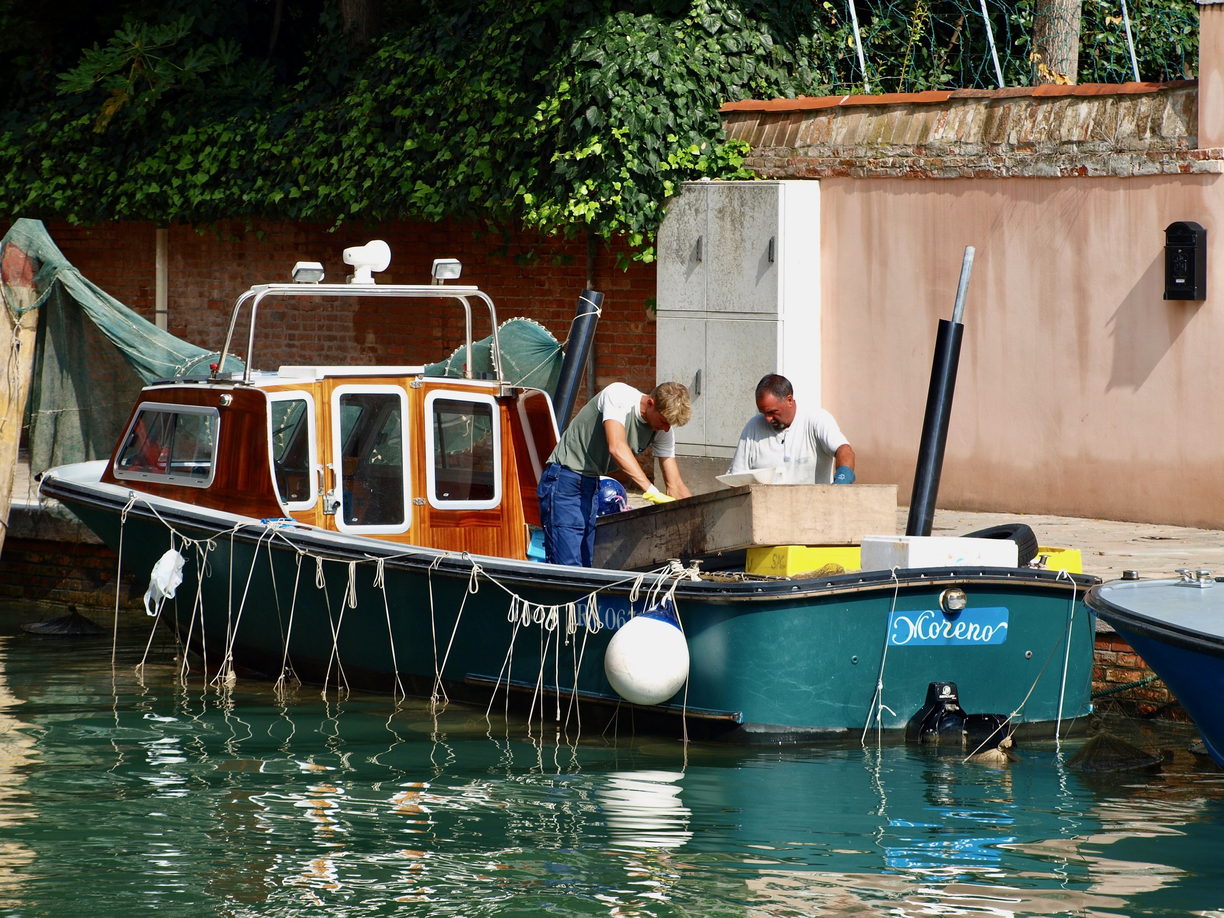 Deux hommes travaillent sur une petite embarcation à quai, avec un bateau nommé 'Morenor' flottant sur l'eau calme, entourée de murets en briques et de feuillages verts.