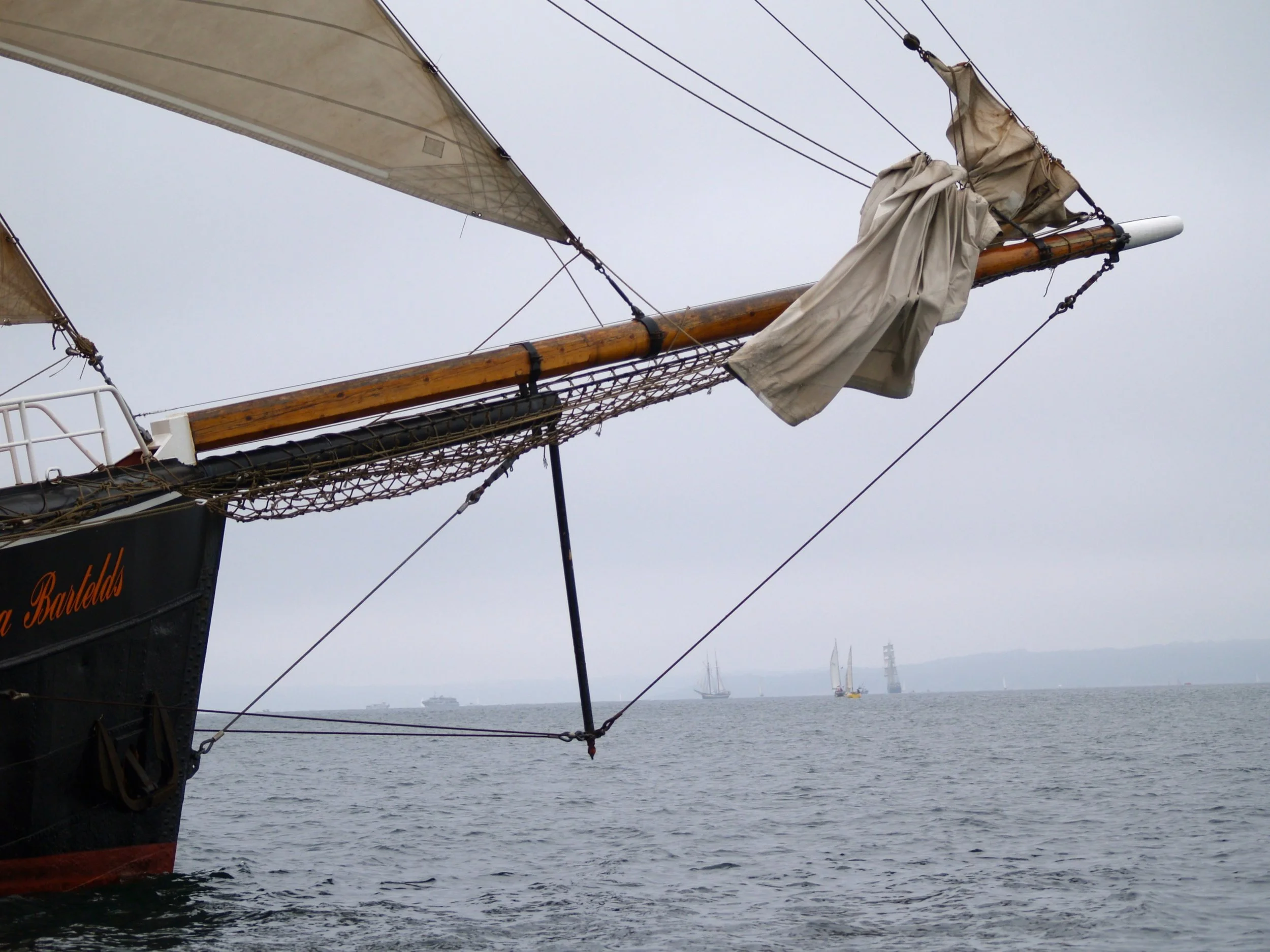Proue d'un vieux navire à voiles avec une voile enroulée, vue sur la mer avec plusieurs autres navires au loin sous un ciel nuageux.