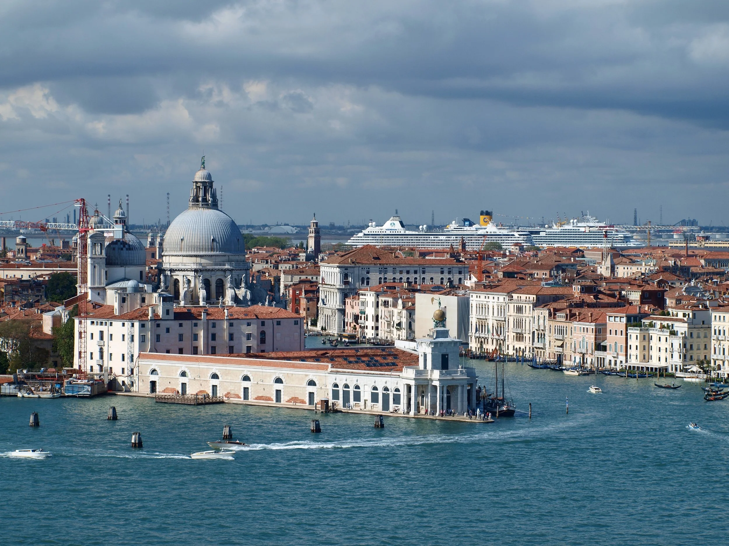 Vue sur le front de mer de Venise avec des bâtiments historiques, des bateaux naviguant sur l'eau, un grand navire de croisière au loin, et un ciel nuageux.