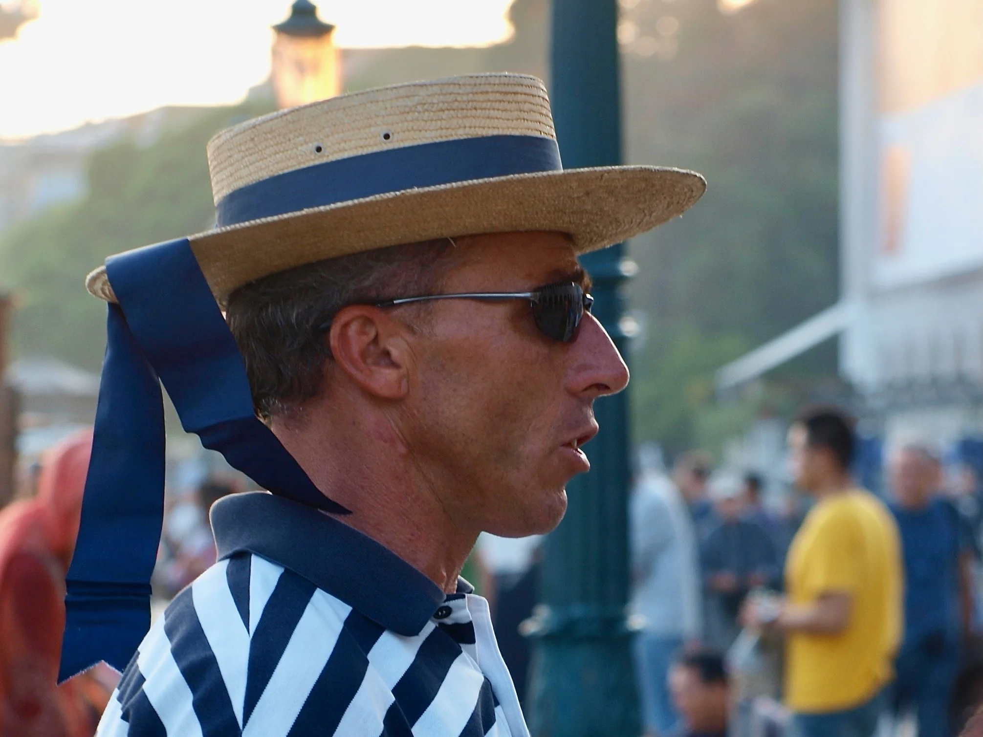 Un homme portant un chapeau de paille beige avec une bande noire, des lunettes de soleil et un maillot de sport à rayures bleues et blanches, regardant vers la droite, lors d'un événement en plein air.