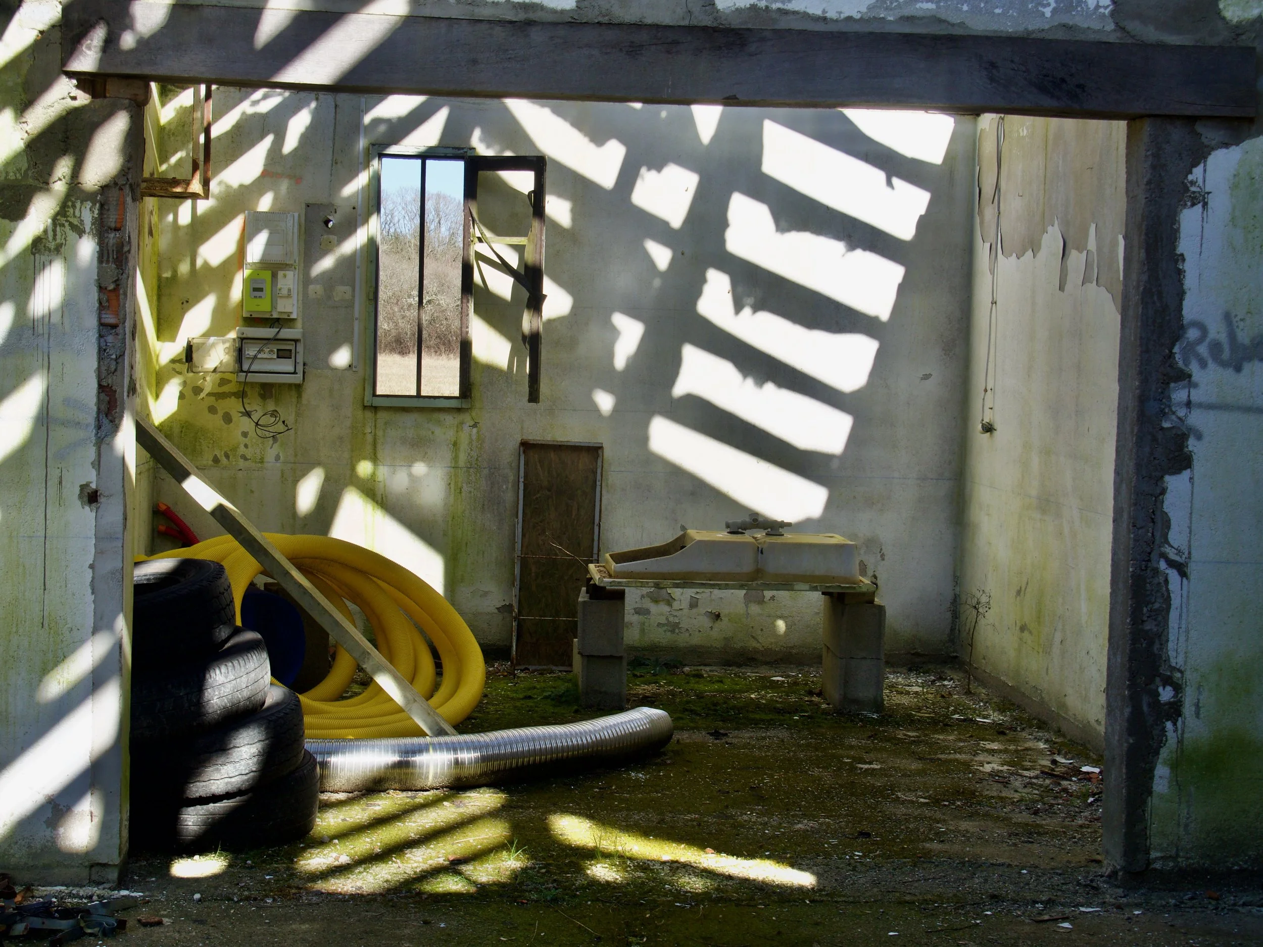 Une structure abandonnée avec des murs fissurés et décolorés, un plafond en béton, diverses tuyaux jaunes et métalliques au sol, et des pneus empilés.