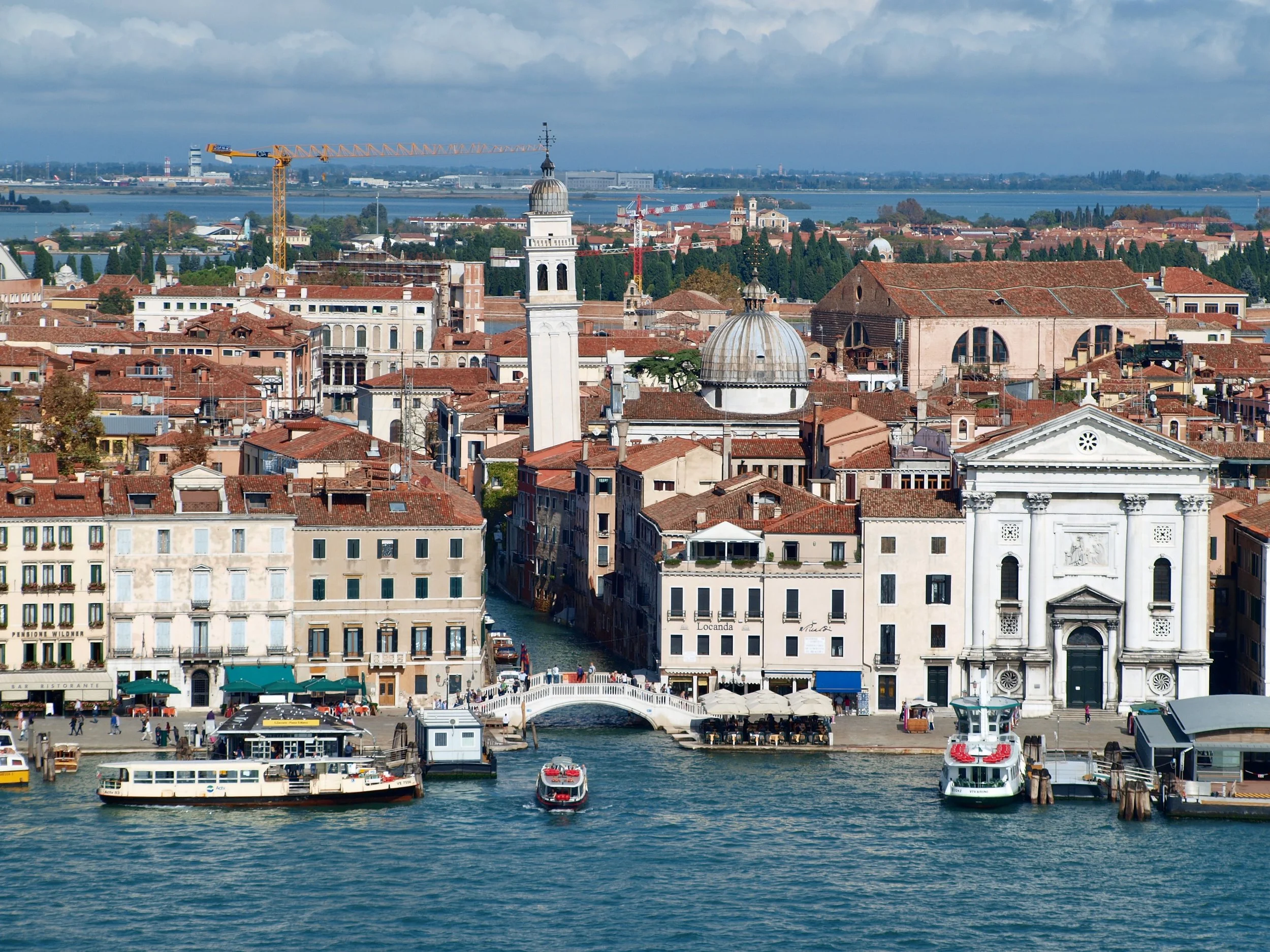 Vue d'une ville italienne avec des bâtiments historiques et une tour, bordée d'une rivière ou d'un canal avec des bateaux.