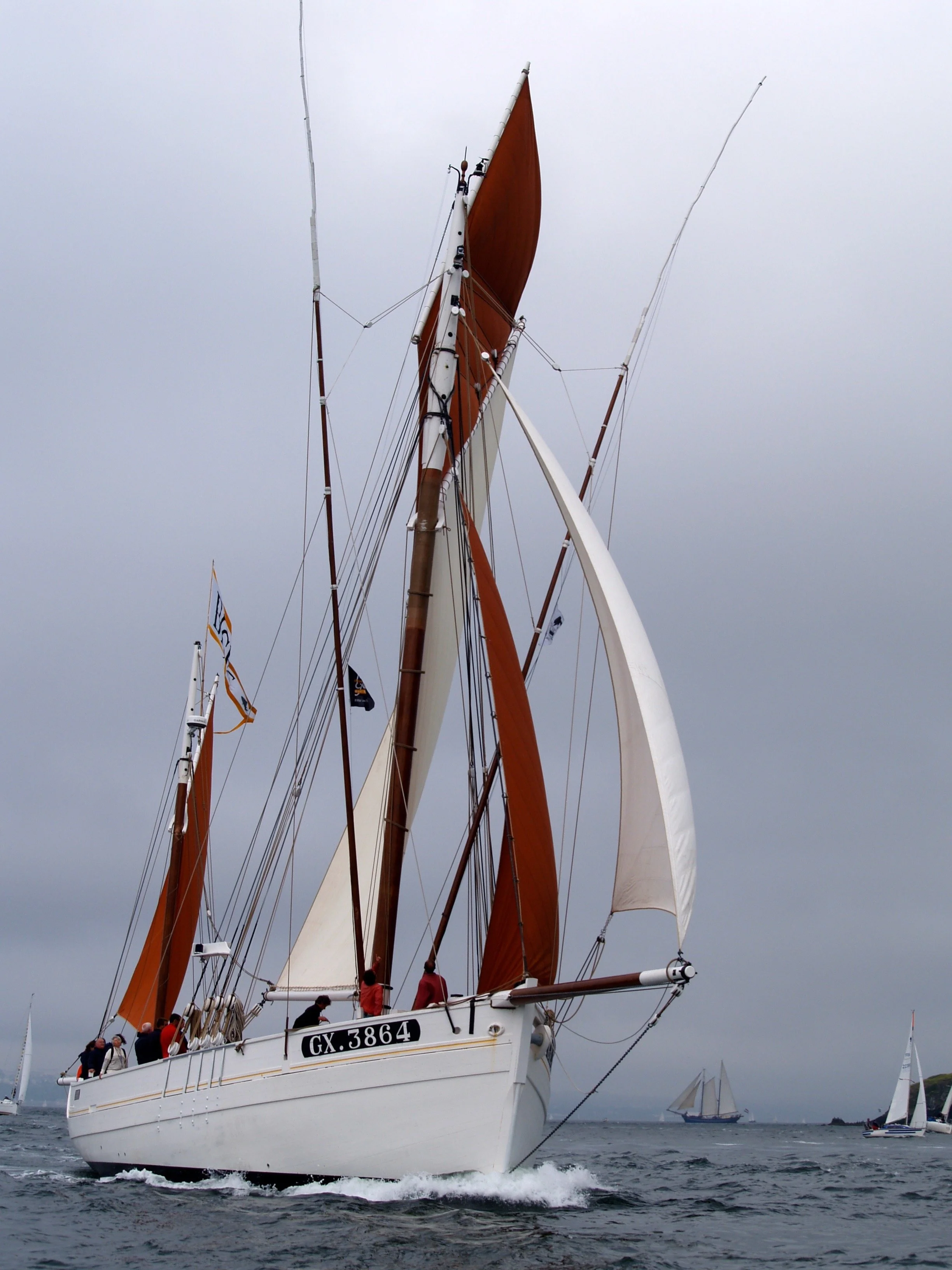 Un voilier blanc avec des voiles en bois orange navigue sur l'eau par temps nuageux, plusieurs autres voiliers en arrière-plan.