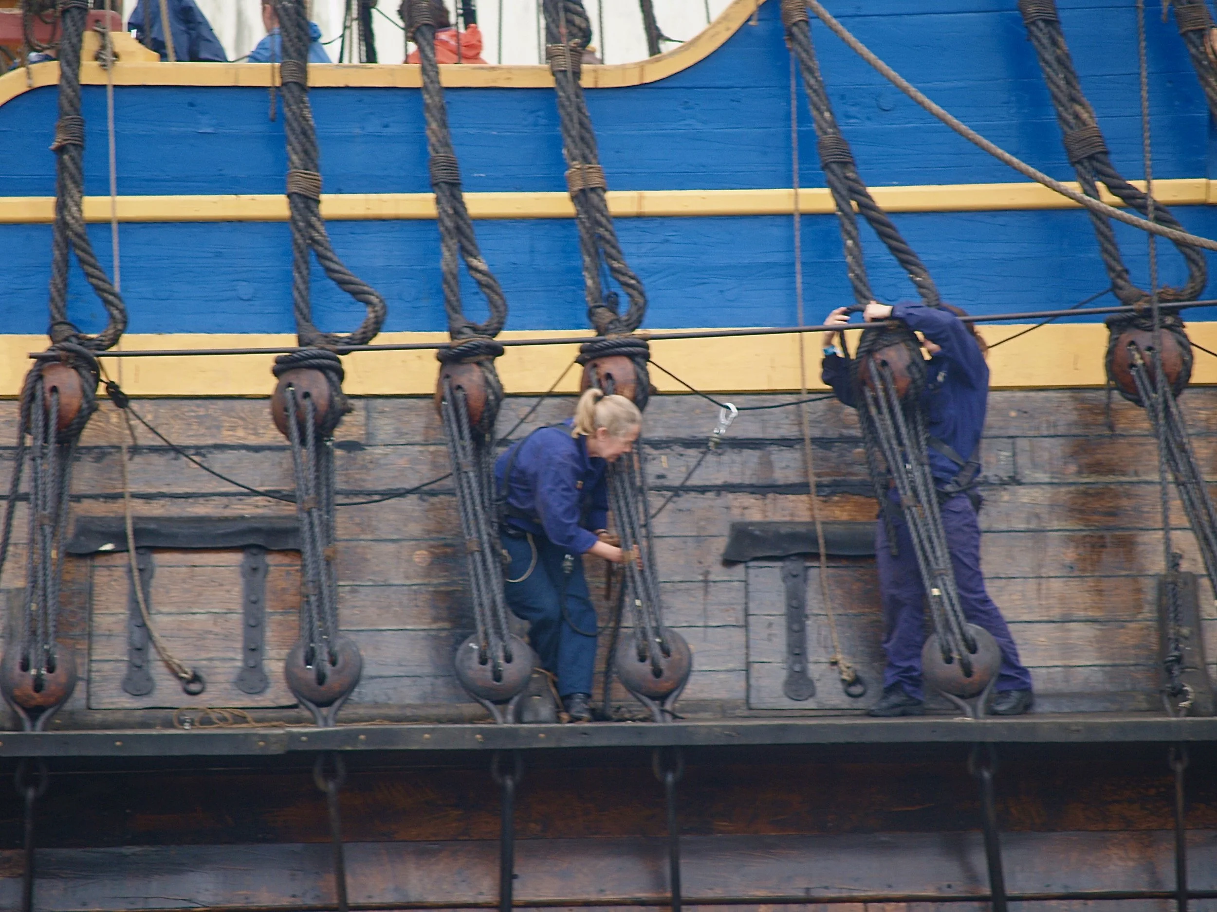 Personnes habillées en bleu travaillant sur un bateau en bois, avec de longues cordes suspendues et des roulettes en bois.