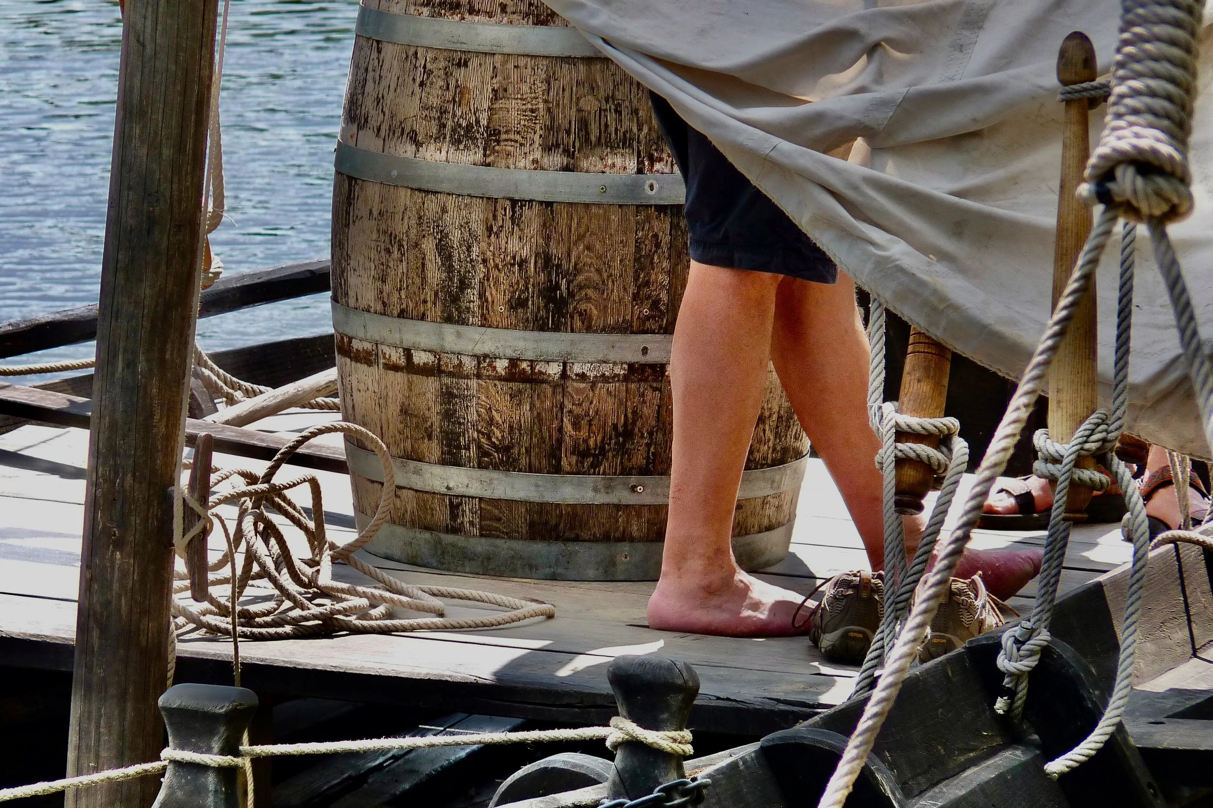 Un pont en bois au bord de l'eau, avec une personne debout portant un pantalon noir et une veste claire, entourée de cordages enroulés.