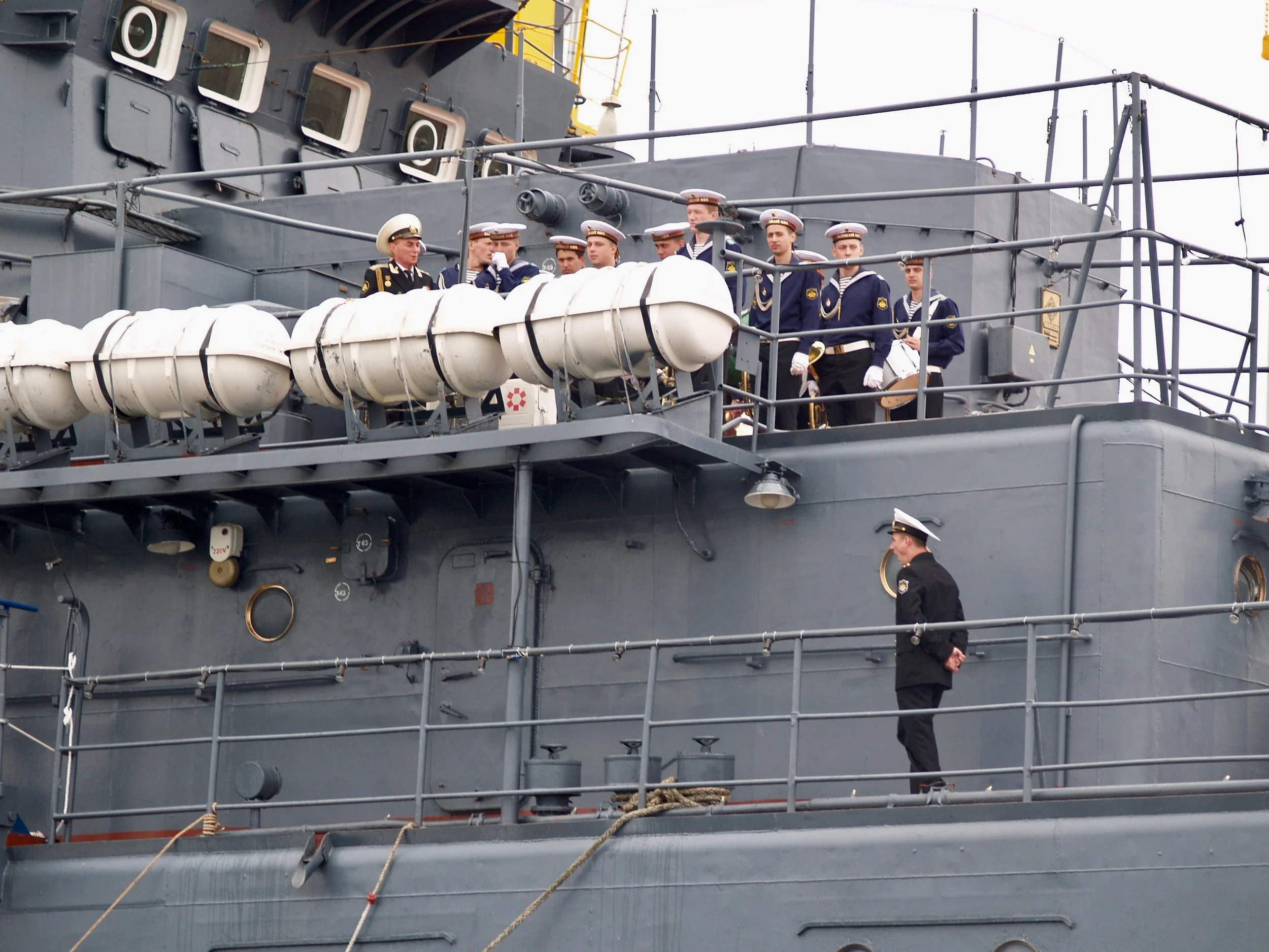 Marins militaires en uniforme sur le pont d'un bateau de guerre, avec une partie de la coque visible et des équipements de bord.