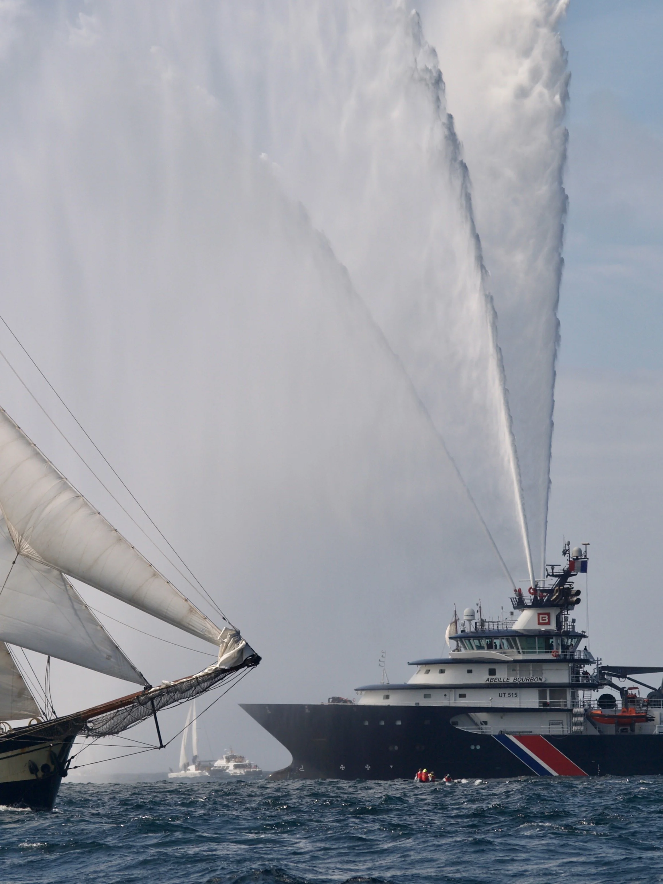 Un navire de la marine effectuant un large jet d'eau au-dessus de la mer, avec un bateau de pêche en premier plan. Le ciel est chargé de nuages gris.