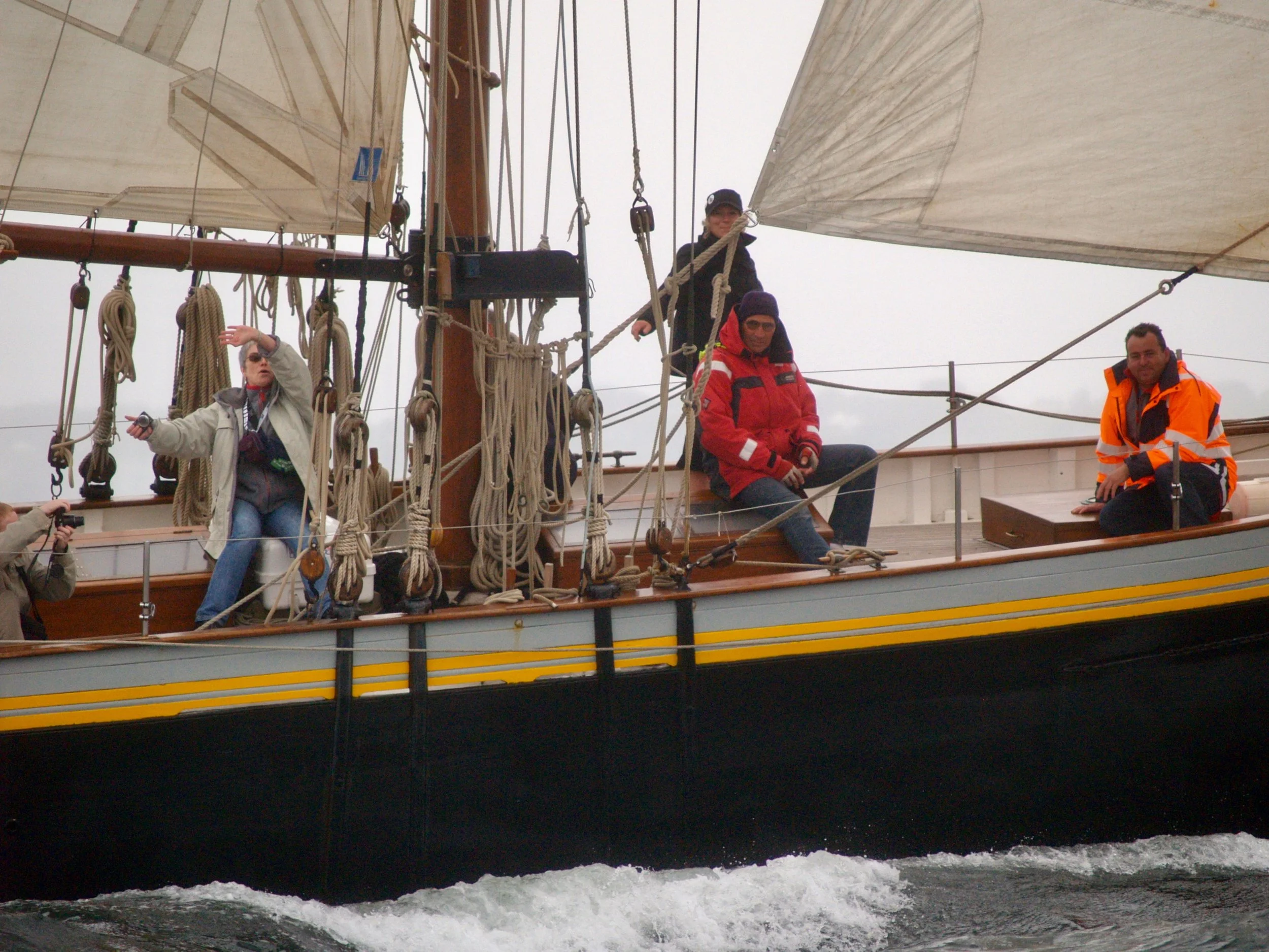 Groupe de trois personnes à bord d'un navire en mer par temps nuageux, avec des cordages, un mât et une voile visibles.