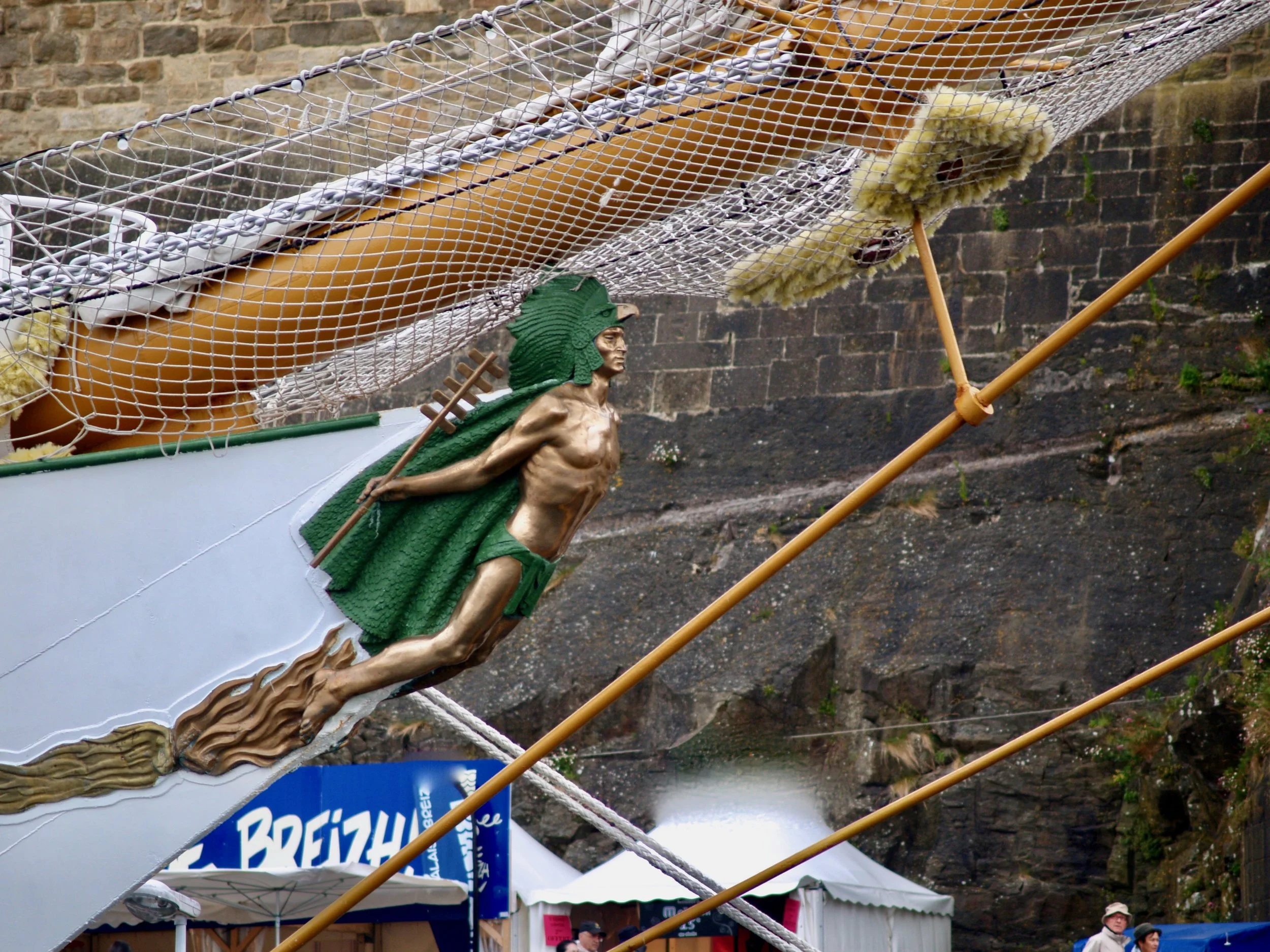 Une représentation artistique d'un héros mythologique, avec une peau de serpent et un casque ailé, tiré d'une affiche ou d'une décoration liée à la Bretagne. En dessous, il y a des tentes ou des stands de marché et des personnes.