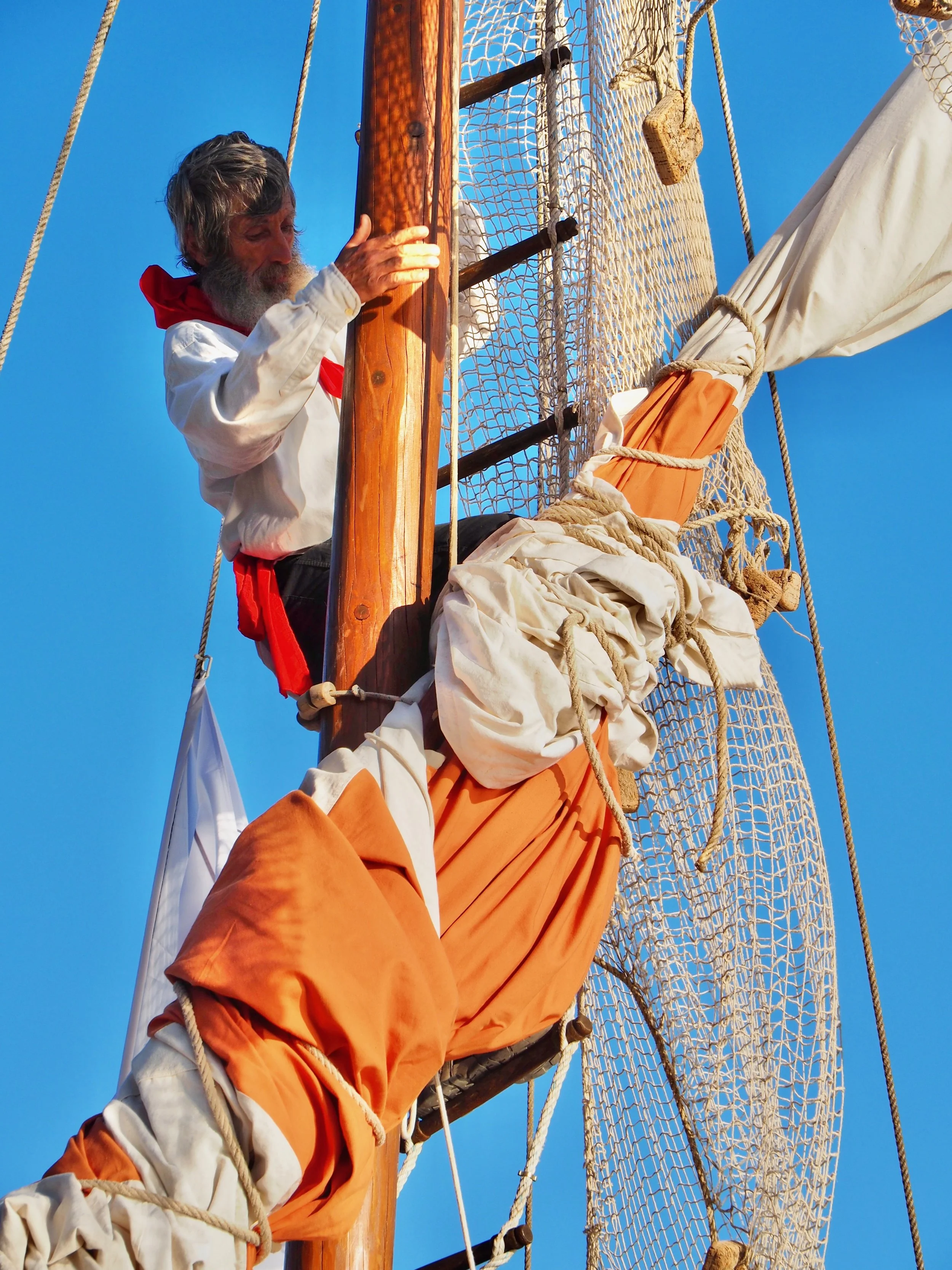Un homme âgé avec une barbe grise, portant une chemise blanche et un foulard rouge, se tient sur le mât d'un voilier, serrant la barre en bois. Des cordages et une voile orange sont visibles, avec un ciel bleu clair en arrière-plan.