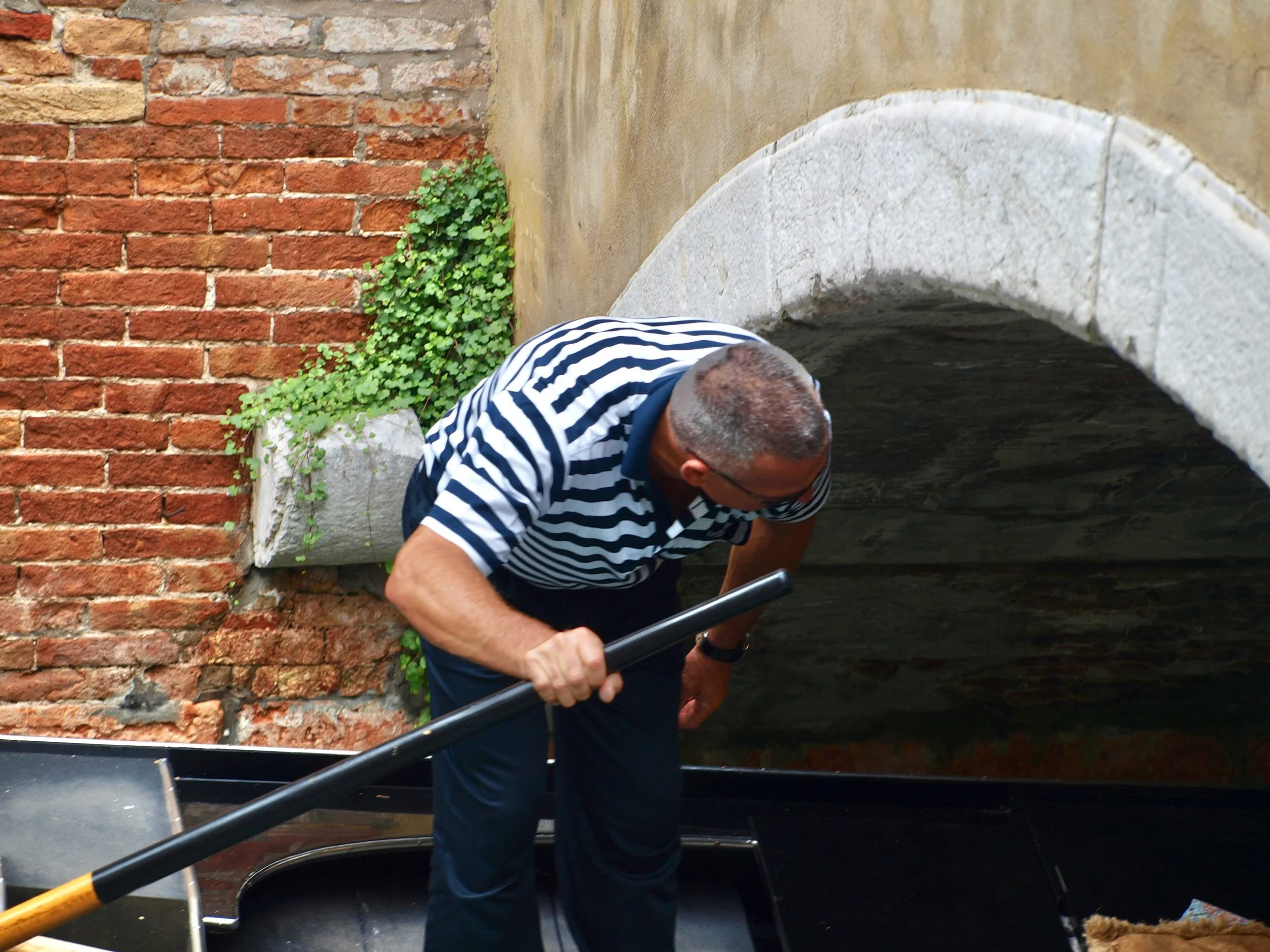 Un homme portant un pull à rayures noires et blanches, avec des cheveux gris, se penche en avant près d'un pont en pierre et d'un mur en briques rouges, sous un ciel nuageux.