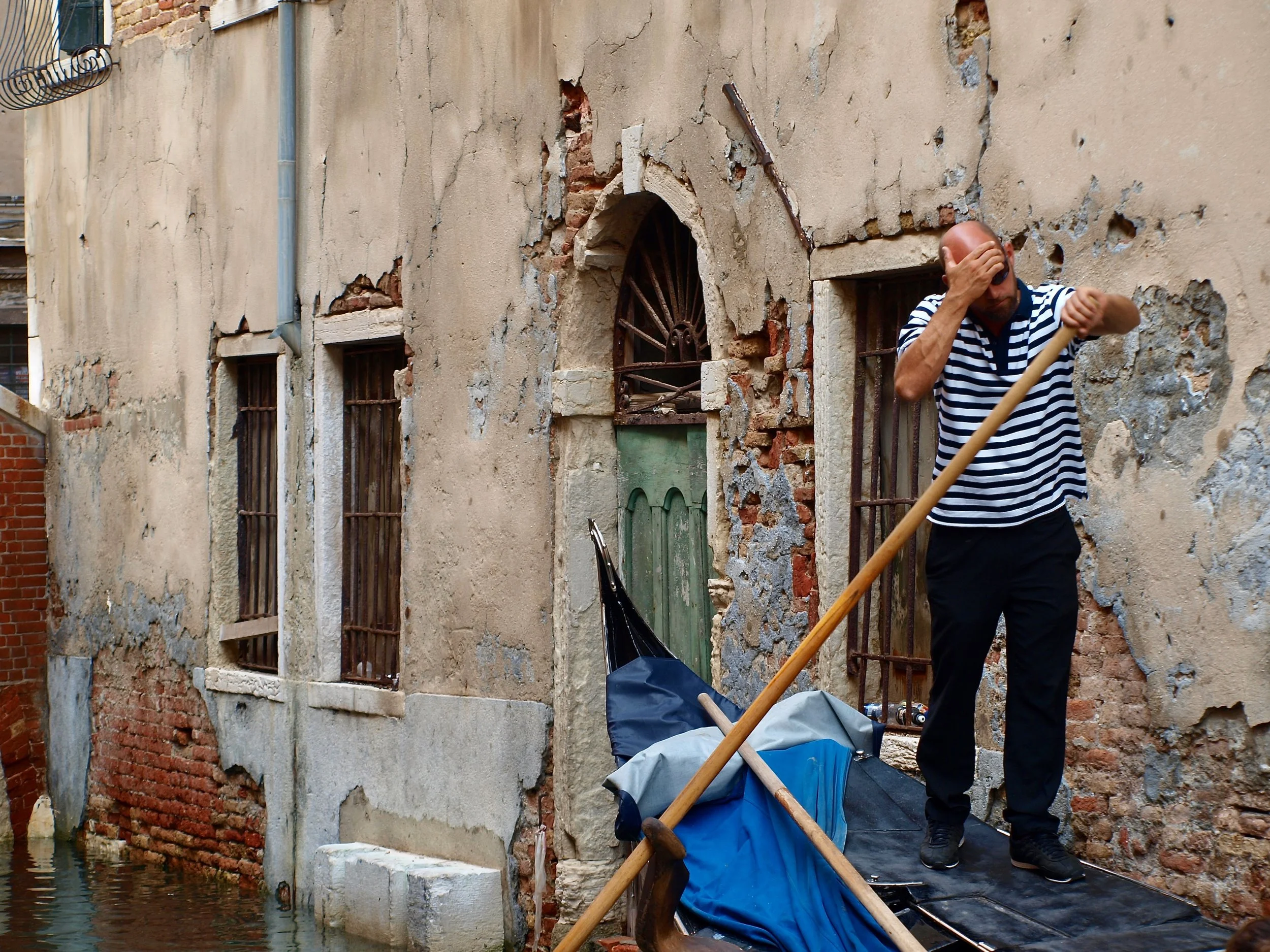 Un homme en marinière est en train de réparer ou restaurer une façade en pierre et brique d'un bâtiment ancien le long d'un canal.