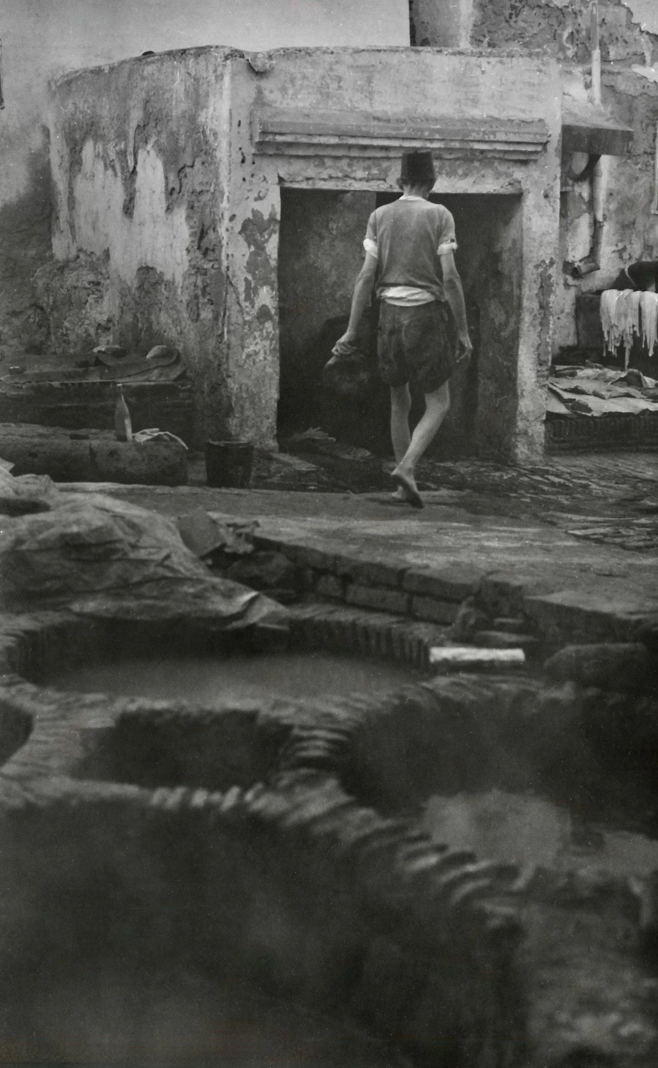 Un jeune garçon marche entre des ruines ou un espace en ruine, avec des murs en pierre ou en béton abîmés, dans un environnement dégradé. L'image est en noir et blanc.