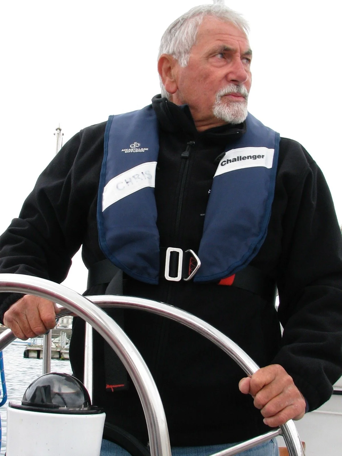 Un homme âgé avec des cheveux gris et une barbe blanche, portant un gilet de sauvetage bleu avec une étiquette indiquant le prénom 'Chris' et le titre 'Challenger'. Il tient le volant d'un bateau, avec un environnement marin en arrière-plan.