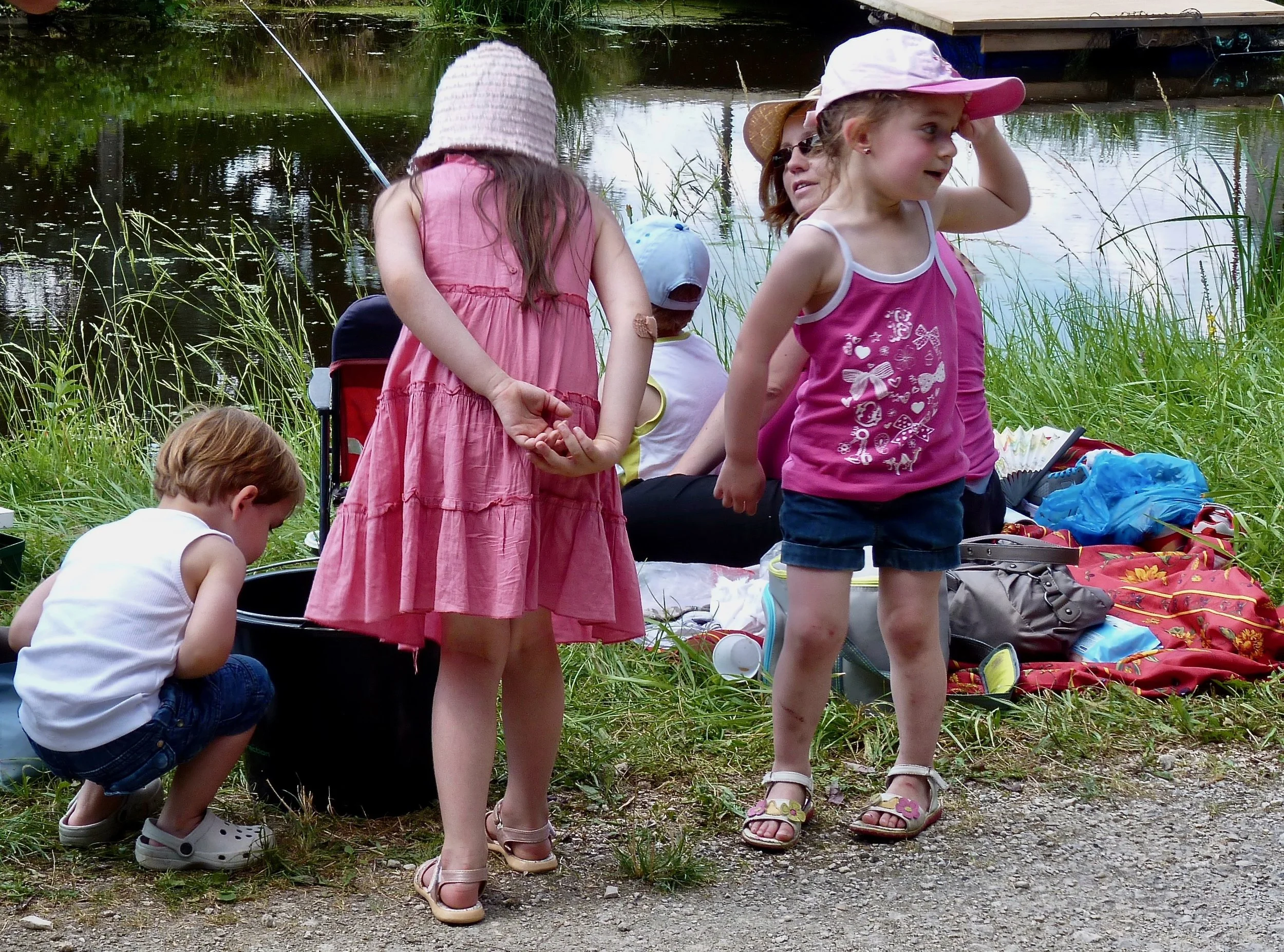 Groupe d'enfants près d'un étang, certains regardant vers l'eau, d'autres observant autour, en vêtements d'été, avec des chapeaux, à l'extérieur dans un environnement naturel.