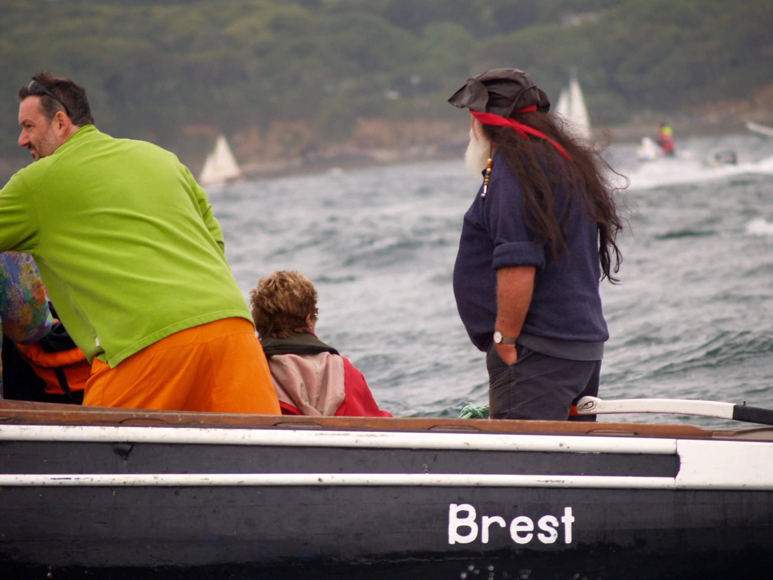 Une image de personnes sur un bateau près de Brest, en Bretagne, avec la mer en arrière-plan. Certaines personnes sont assises ou debout, portant des vêtements colorés et un bandana rouge. Des voiliers sont visibles au loin sur l'eau.