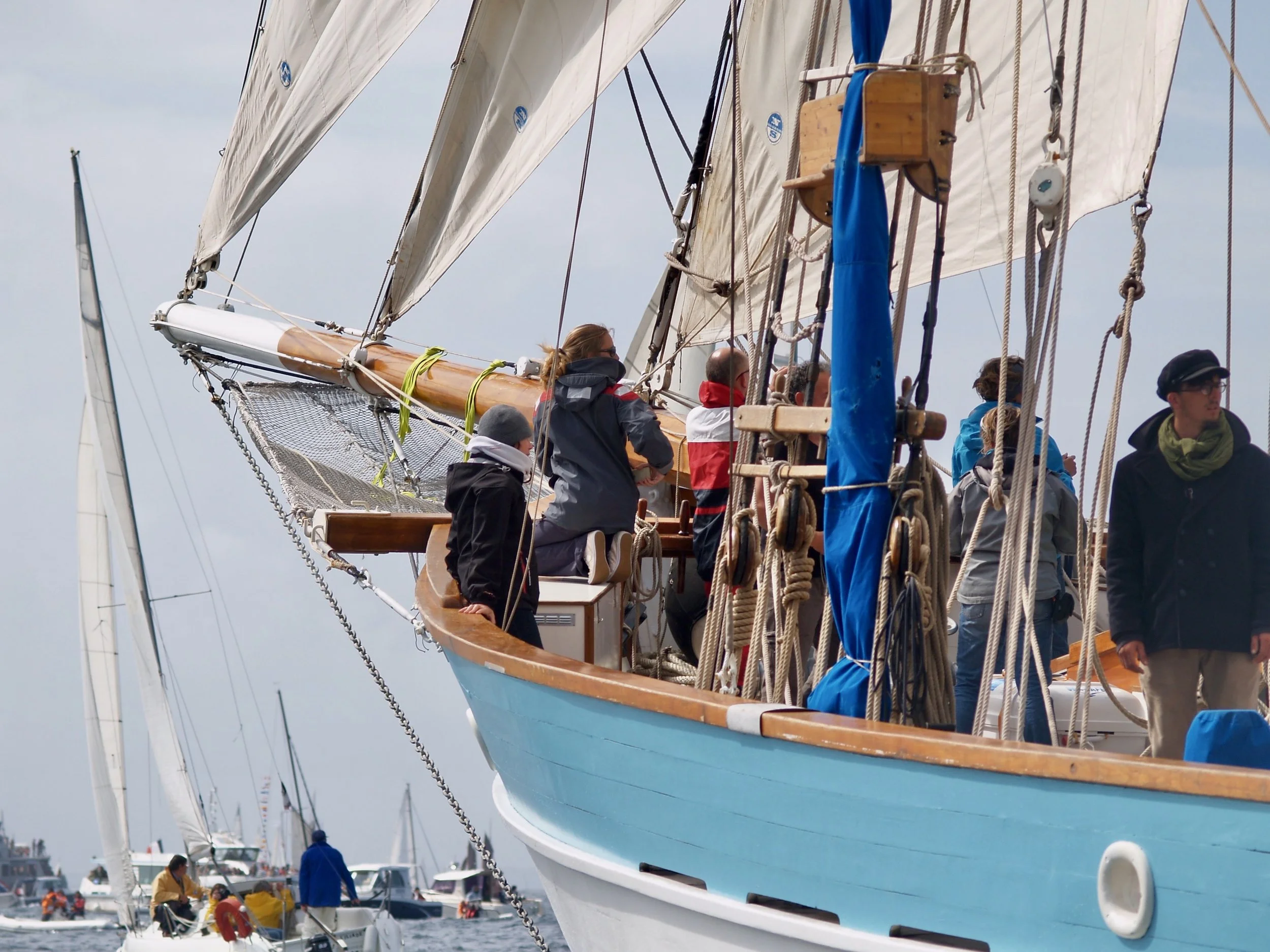 Un voilier avec plusieurs personnes à bord, certains regardant vers l'avant, dans un port avec d'autres bateaux, à l'extérieur lors d'une journée nuageuse.