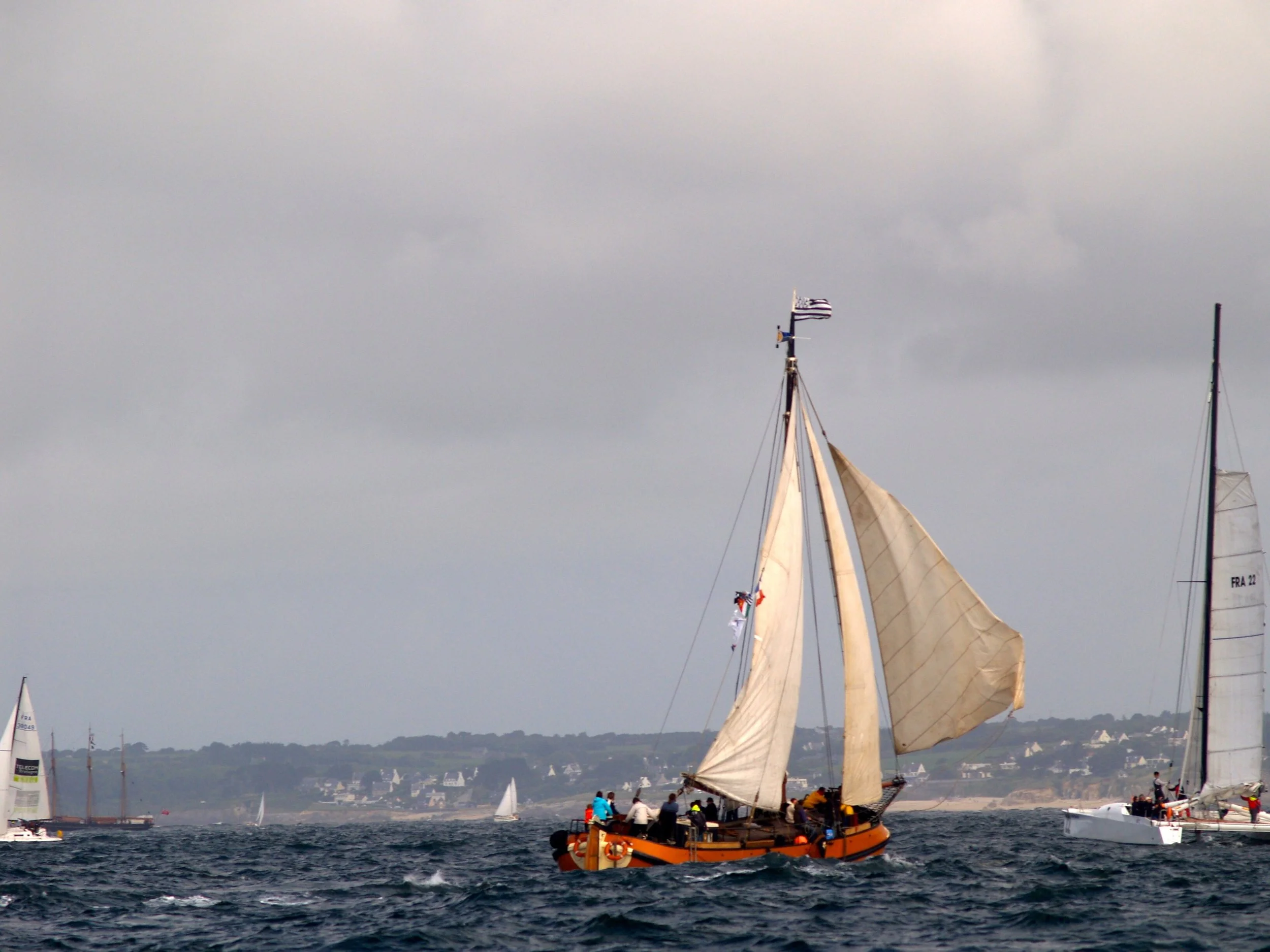 Un voilier en mer avec un drapeau américain et plusieurs autres voiliers en arrière-plan, sous un ciel nuageux.