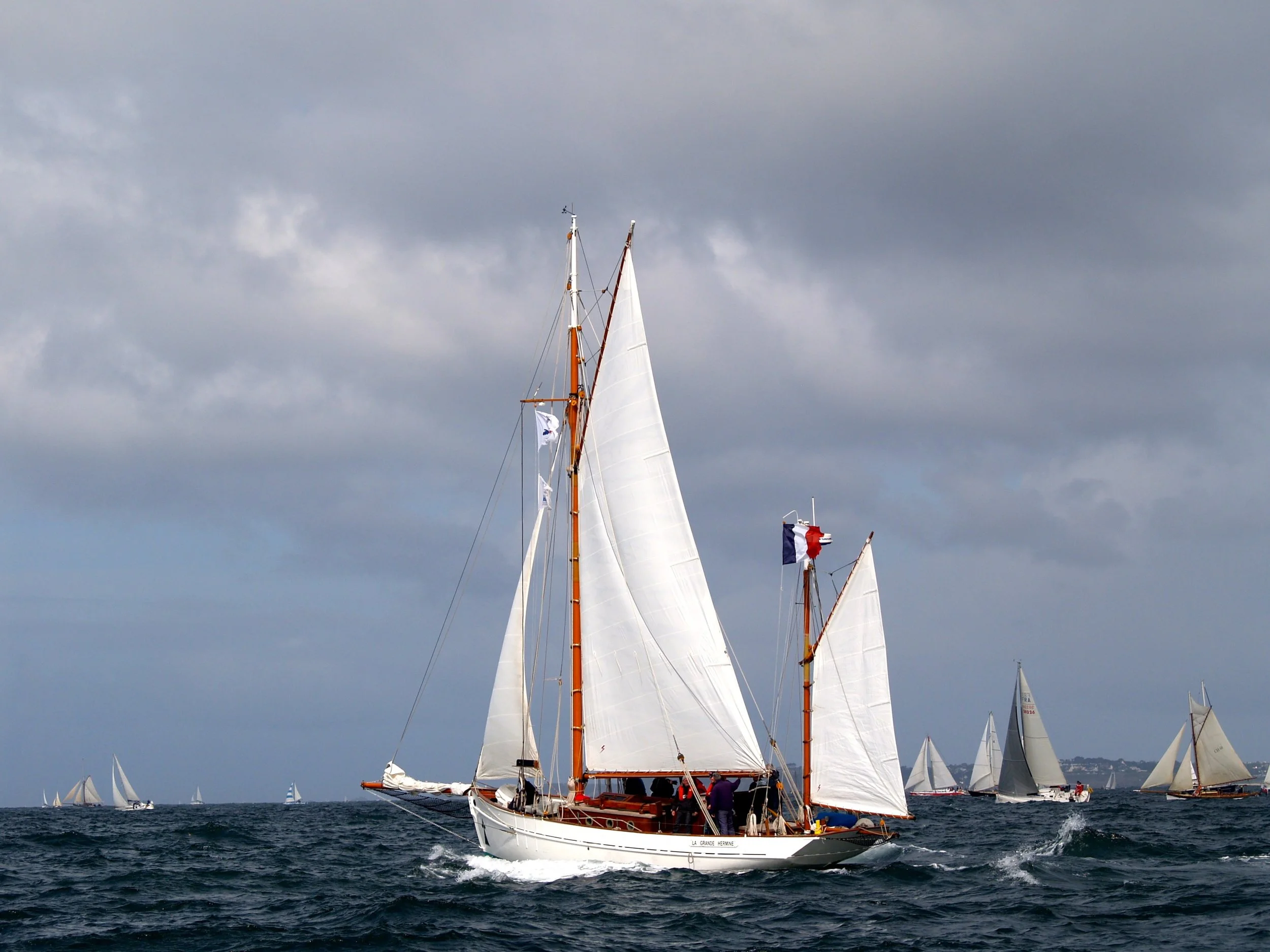 Un voilier navigue sur la mer forte avec plusieurs autres voiliers en arrière-plan sous un ciel nuageux.