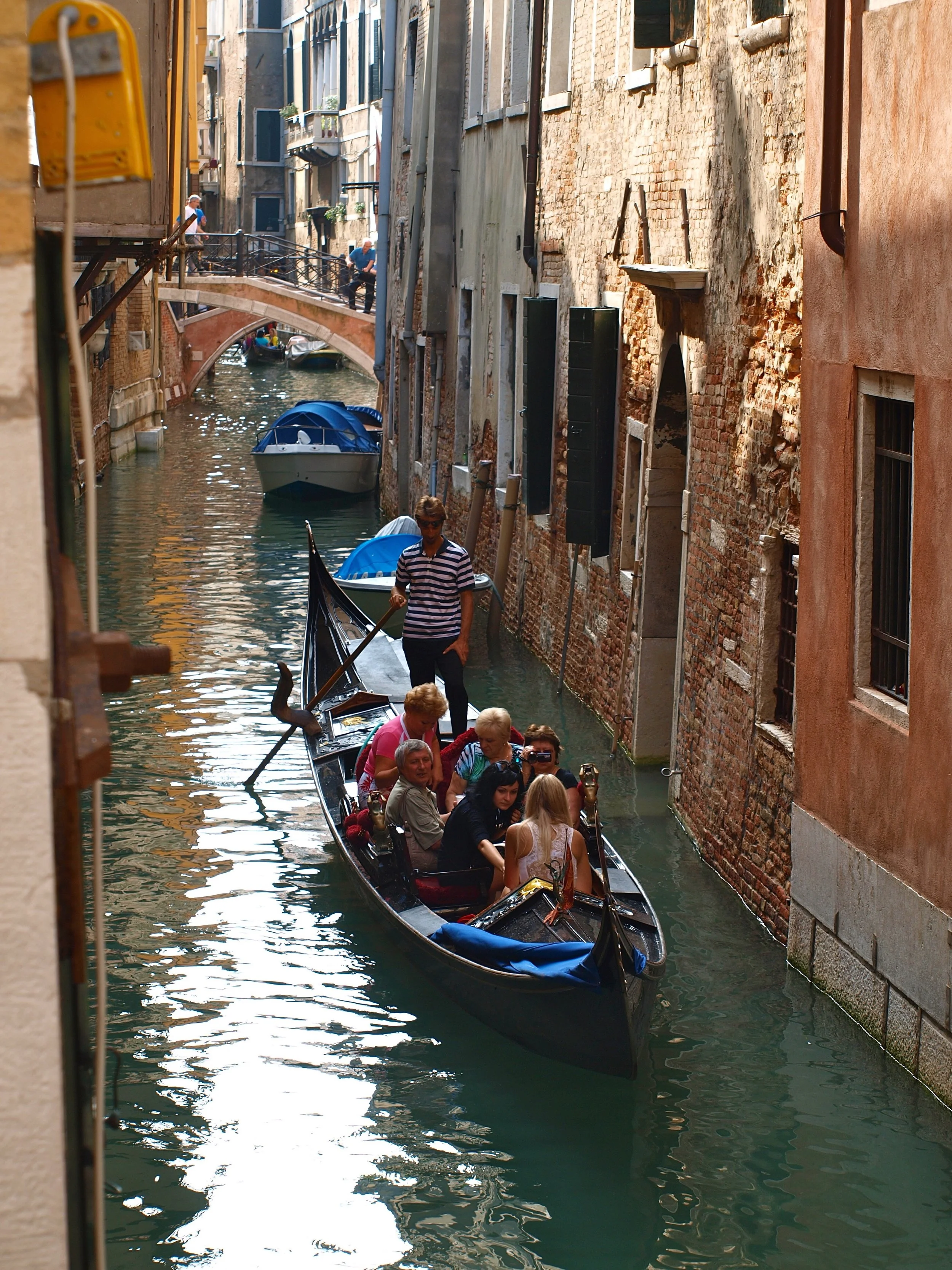 Gondole transportant des touristes dans un canal de Venise, avec des bâtiments anciens en brique sur les côtés, un pont en pierre en arrière-plan, et des personnes assises dans la gondole.