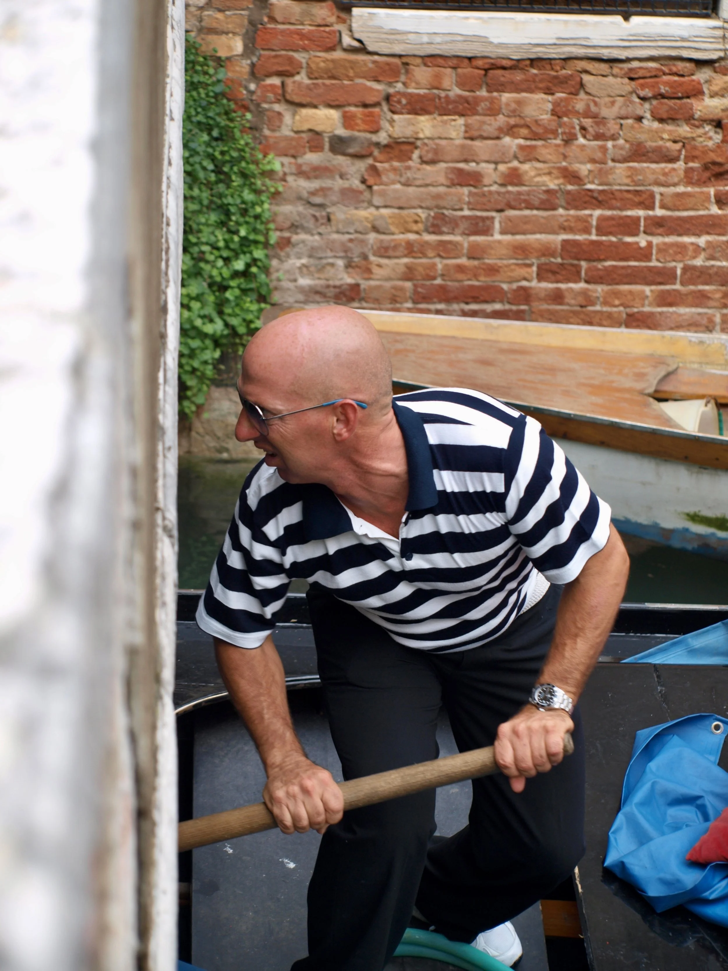 Homme chauve portant des lunettes, en chemise à rayures, en train de travailler avec un outil sur un bateau en bois dans une cour en arrière avec des murs en briques.