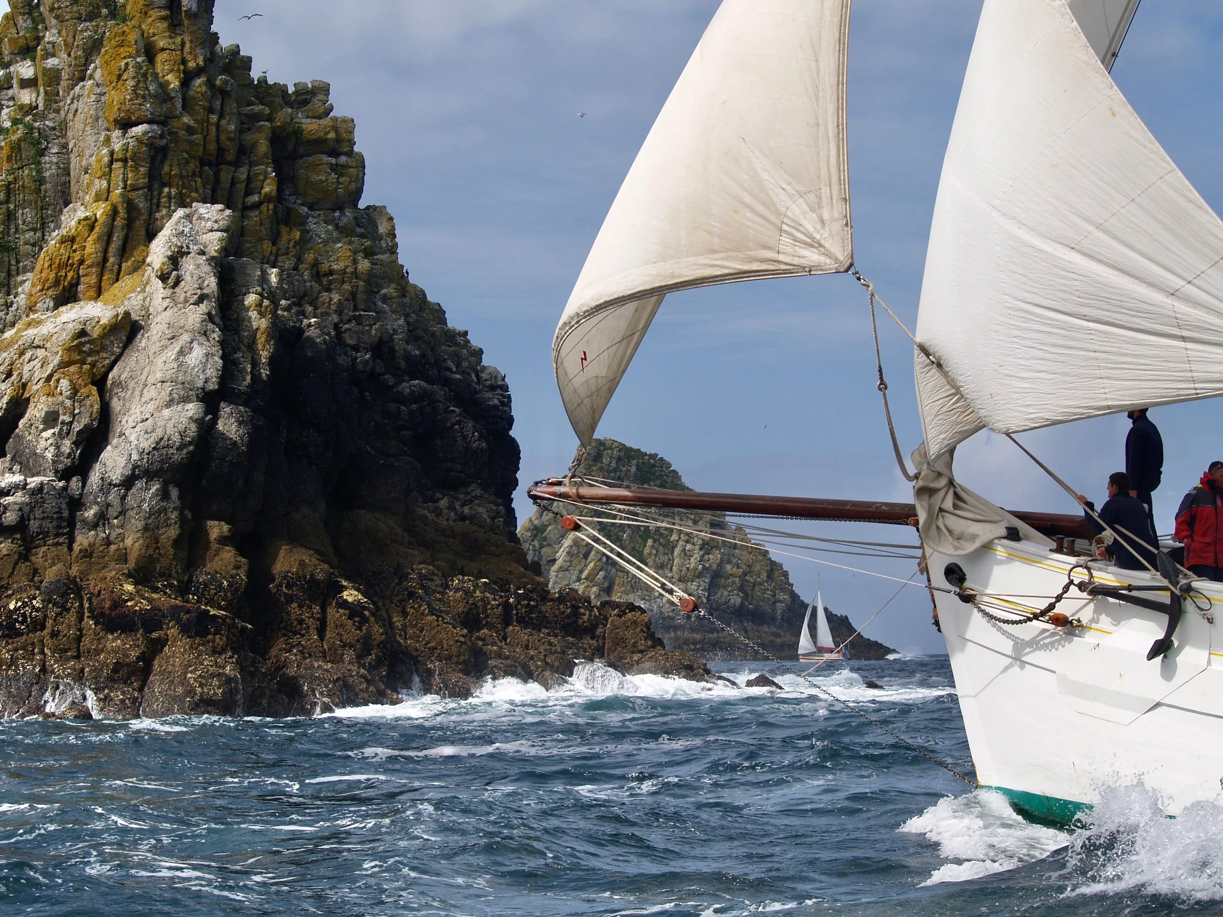 Un voilier avec des voiles blanches navigue près de falaises rocheuses sous un ciel nuageux.