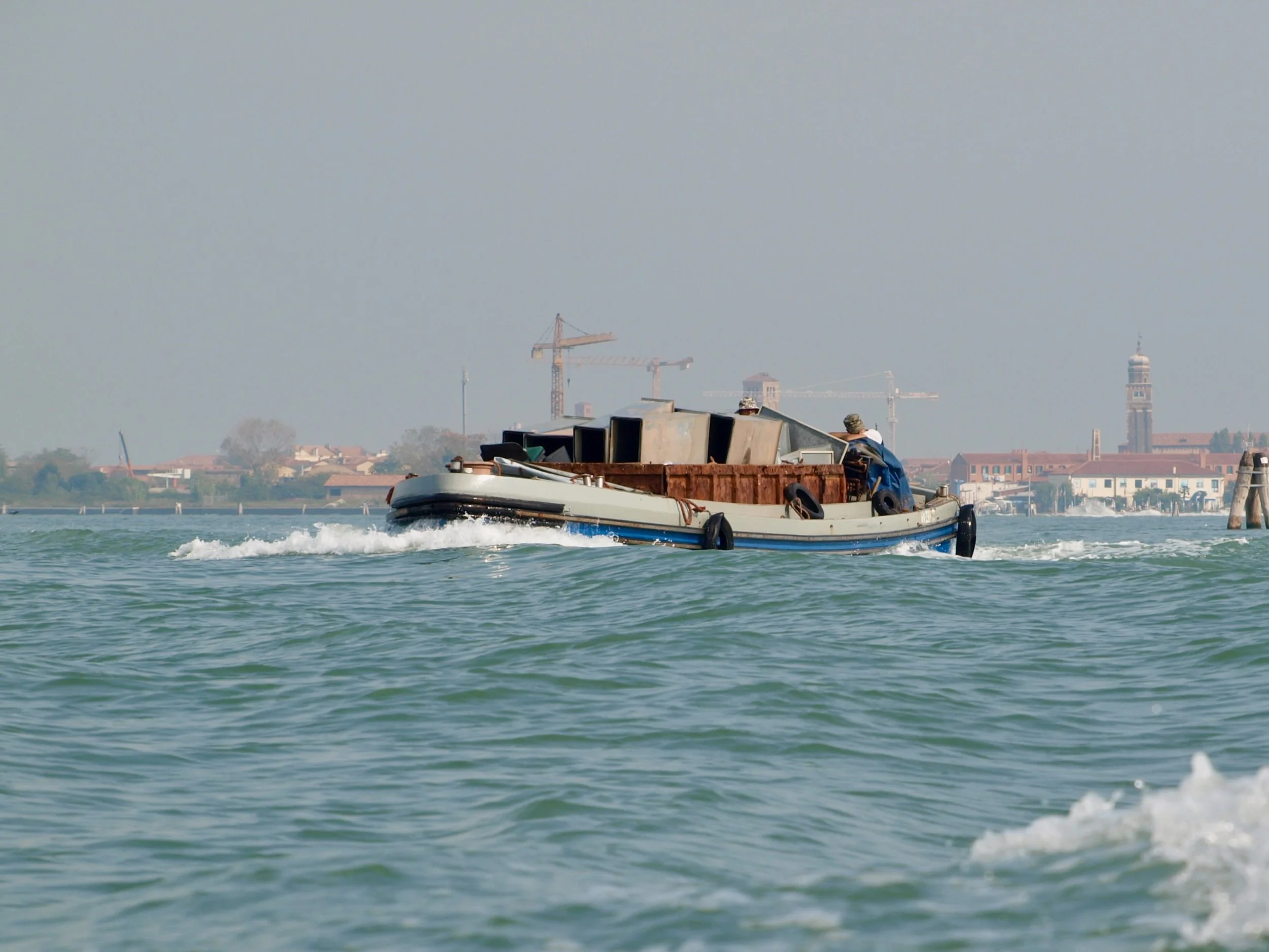 Un bateau navigue sur une rivière ou un lac avec une ville en arrière-plan, comprenant plusieurs grues de construction et un clocher ou une tour. Le ciel est clair et l'eau est légèrement agitée.