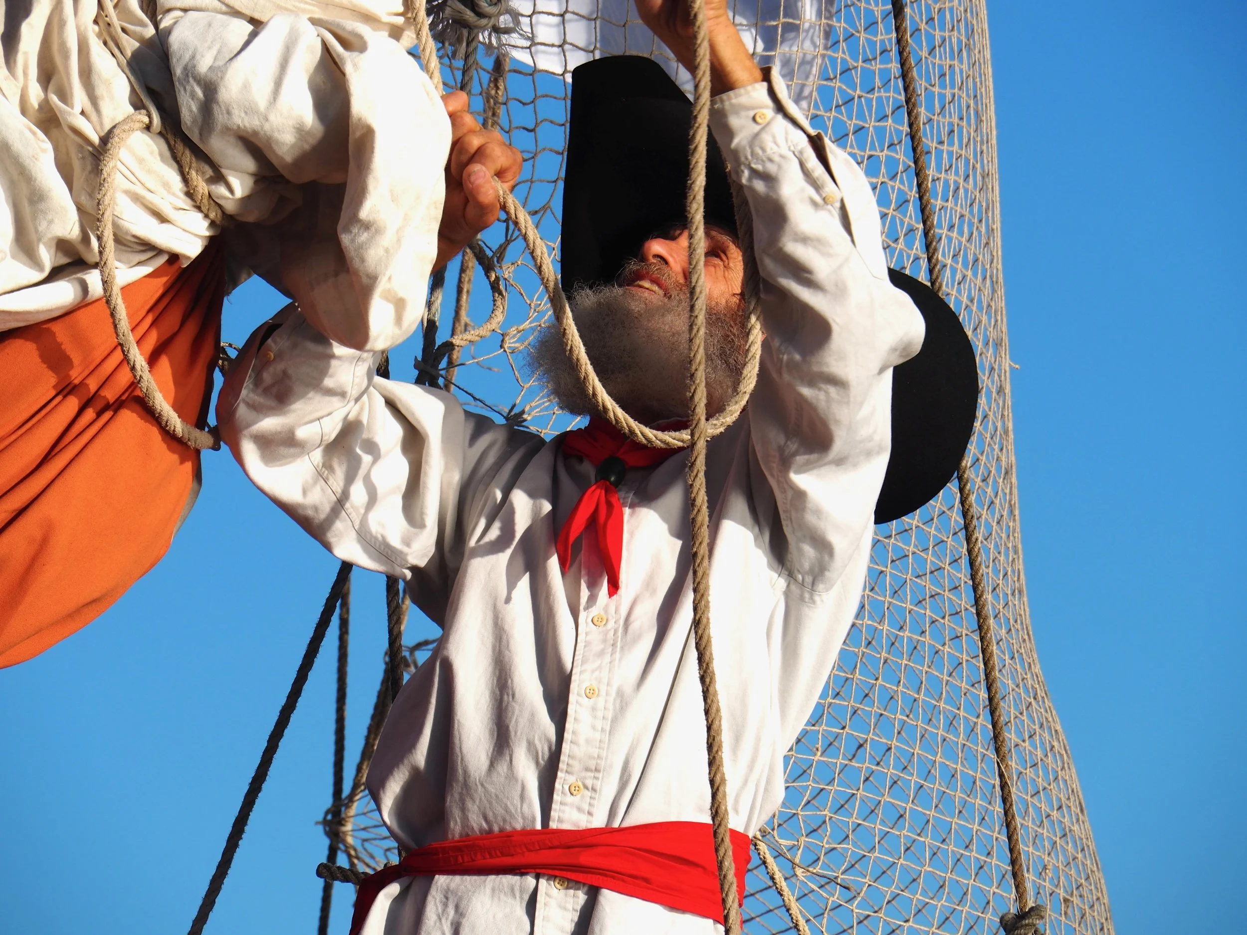 Une personne en costume traditionnel, portant un foulard rouge, grimpe dans une cage sur une montgolfière, avec un ciel bleu en arrière-plan.