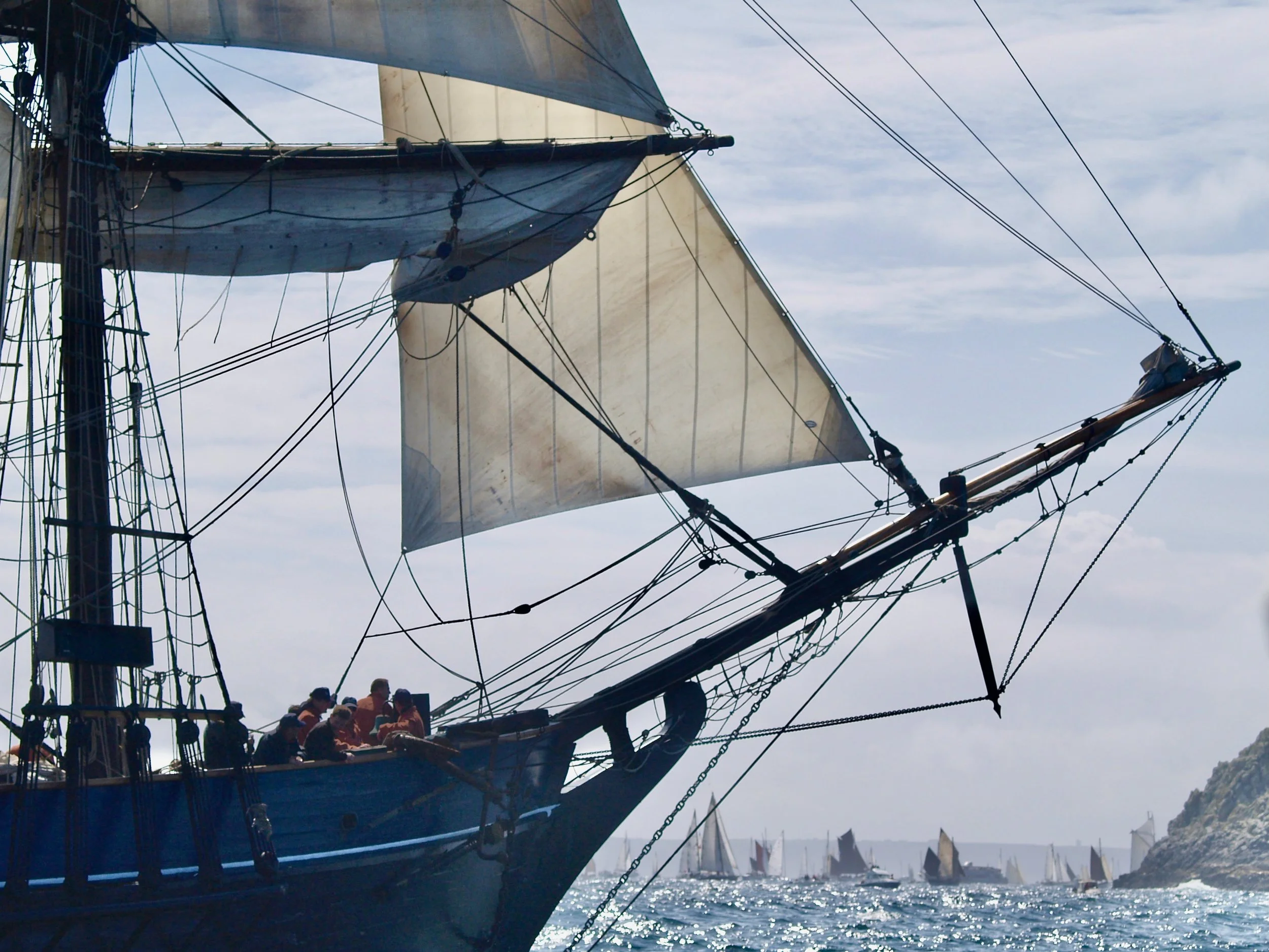 Un grand voilier avec plusieurs mats et voiles, avec des personnes sur le pont, naviguant en mer. D'autres voiliers plus petits sont visibles à l'arrière-plan, avec un ciel partiellement nuageux.