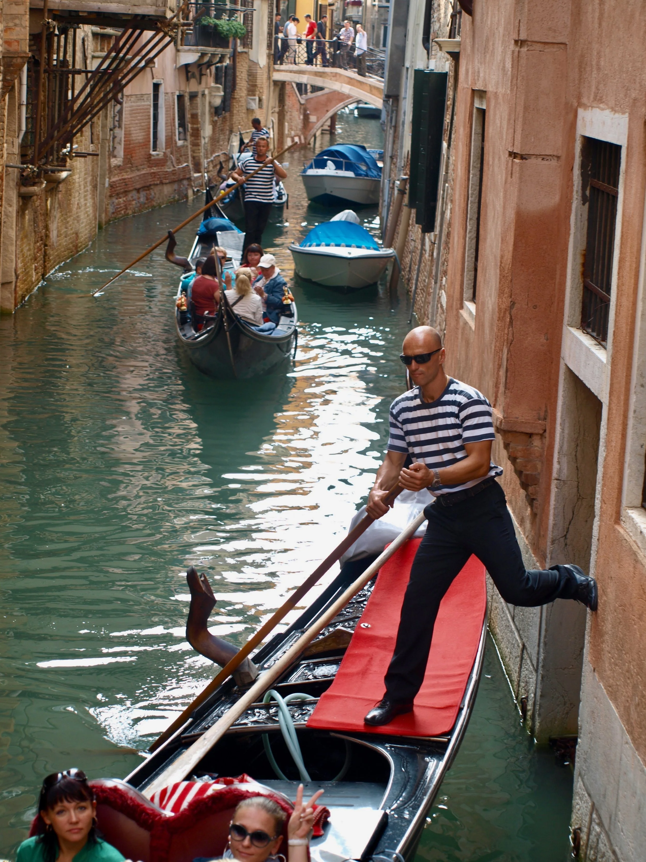 Un gondolier en tenue rayée et lunettes noires navigue dans un canal de Venise, avec des passagers assis dans la gondole. La scène est encadrée par des bâtiments anciens aux murs en pierre et en brique.