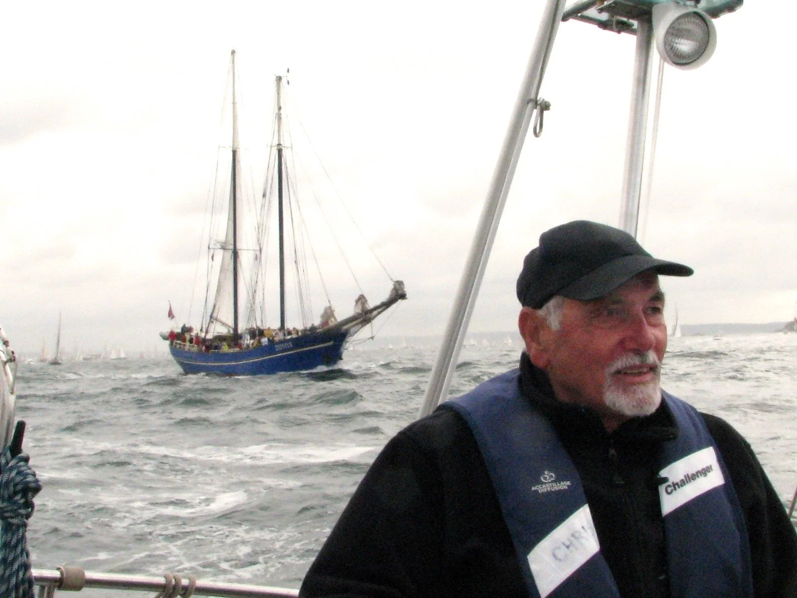 Un homme âgé souriant portant un gilet de vie, en mer, avec un grand voilier à l'arrière-plan par temps nuageux.