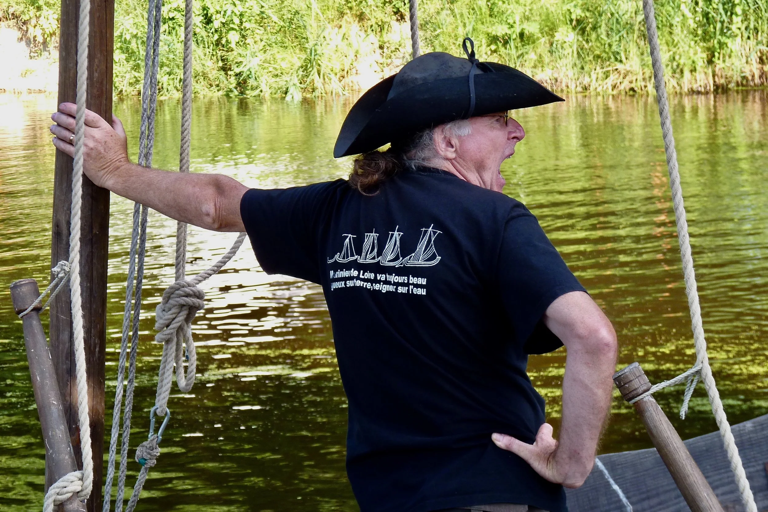 Homme âgé en costume d'époque avec chapeau tricorne, criant, sur un bateau en bois en bord de rivière avec des végétations vertes en arrière-plan.