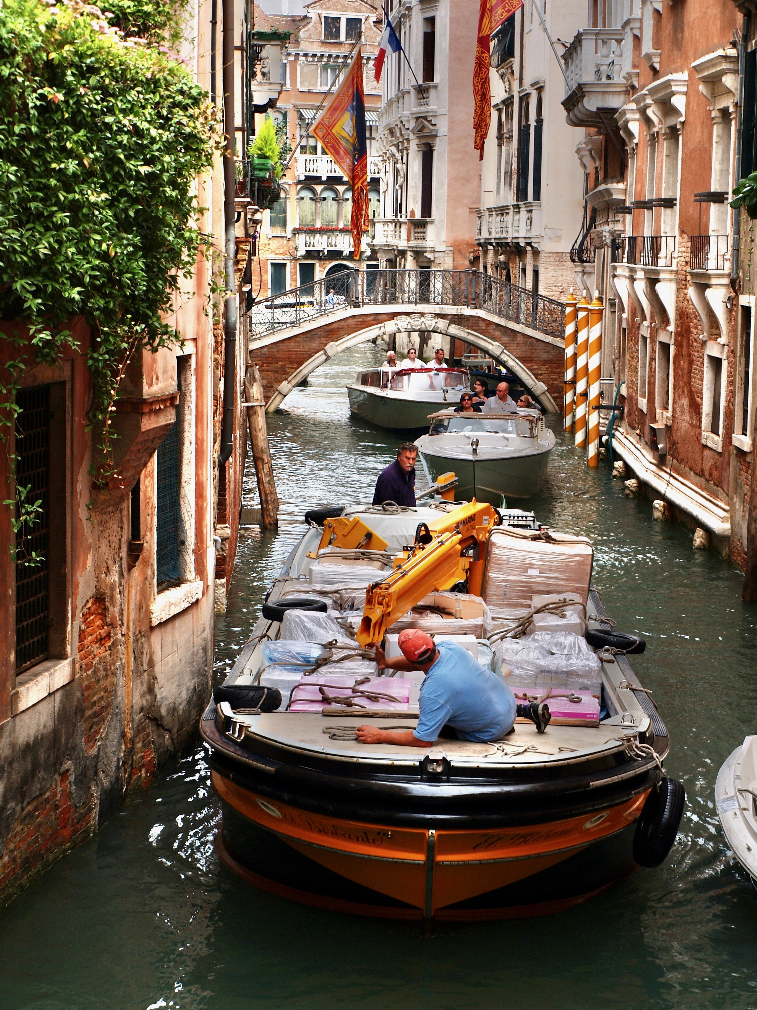 Une scène dans un canal de Venise, avec des bateaux de touristes et des bâtiments historiques. Un homme est sur un bateau chargé de colis ou d'équipements.