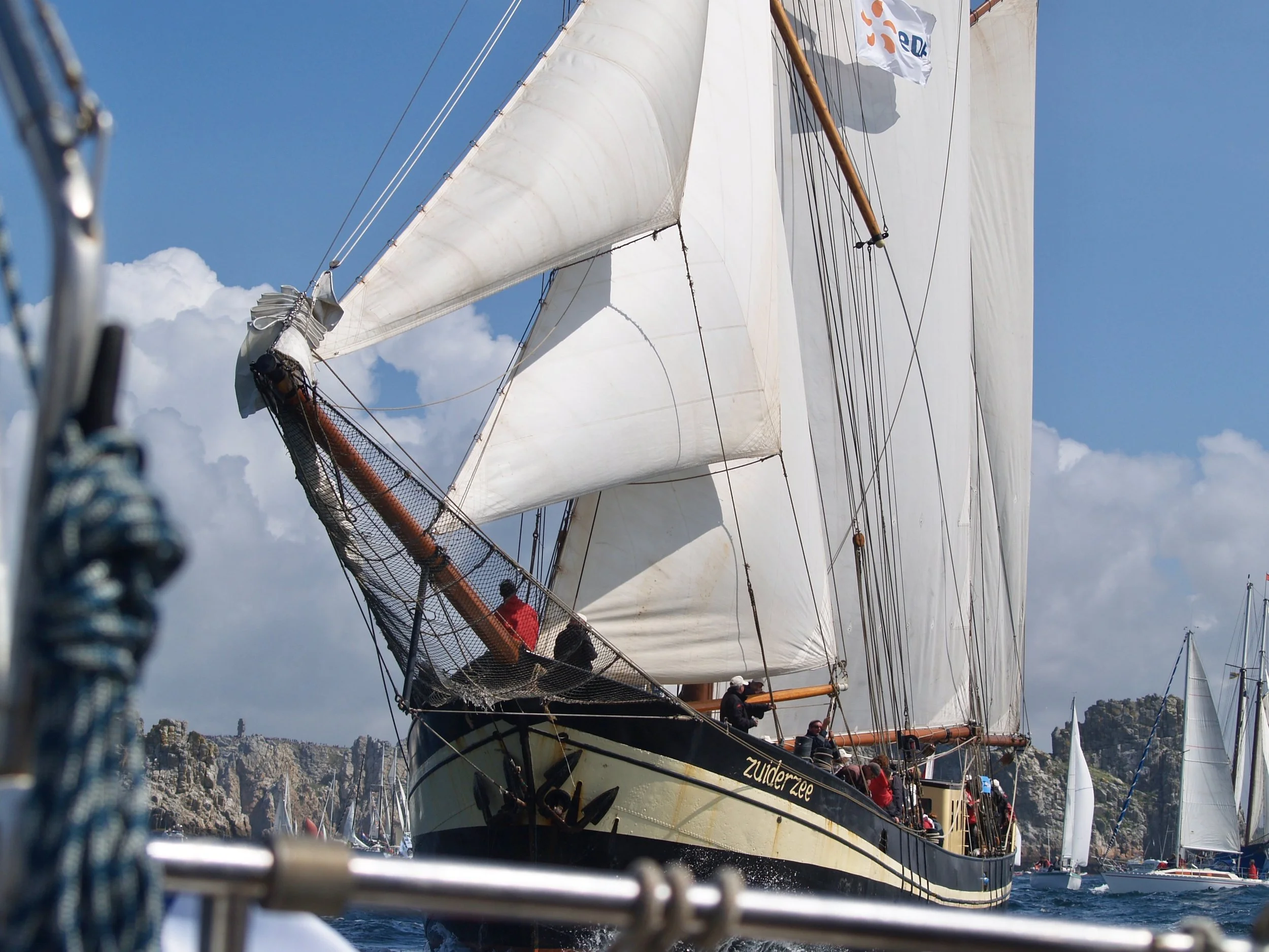 Un grand voilier avec plusieurs voiles blancs est photographié en mer, des personnes sont visibles sur le bateau, avec un paysage rocheux et des bâtiments en arrière-plan.