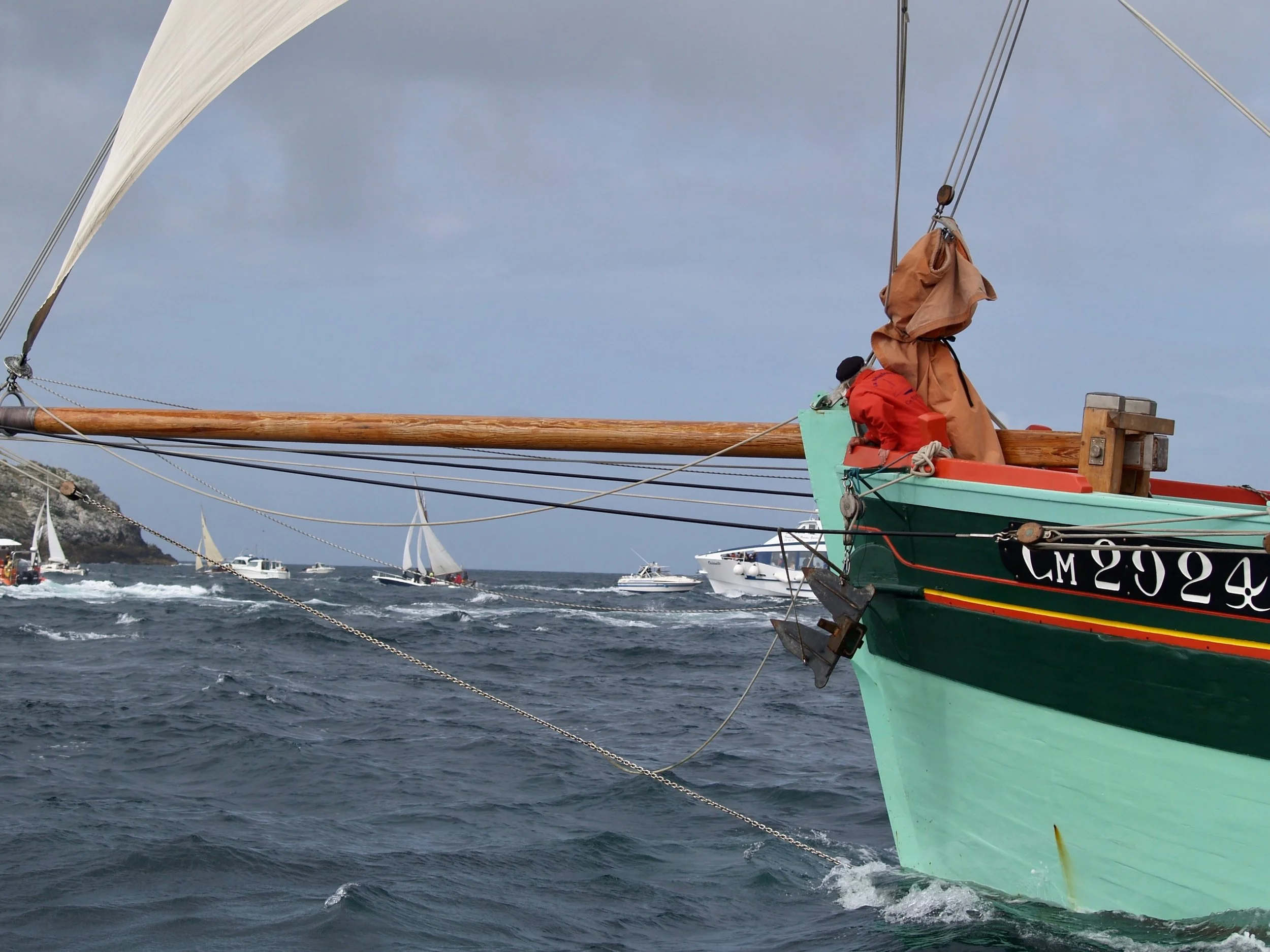 Un voilier coloré avec des voiles blanches navigue en mer avec plusieurs autres barcos et un ciel nuageux en arrière-plan.
