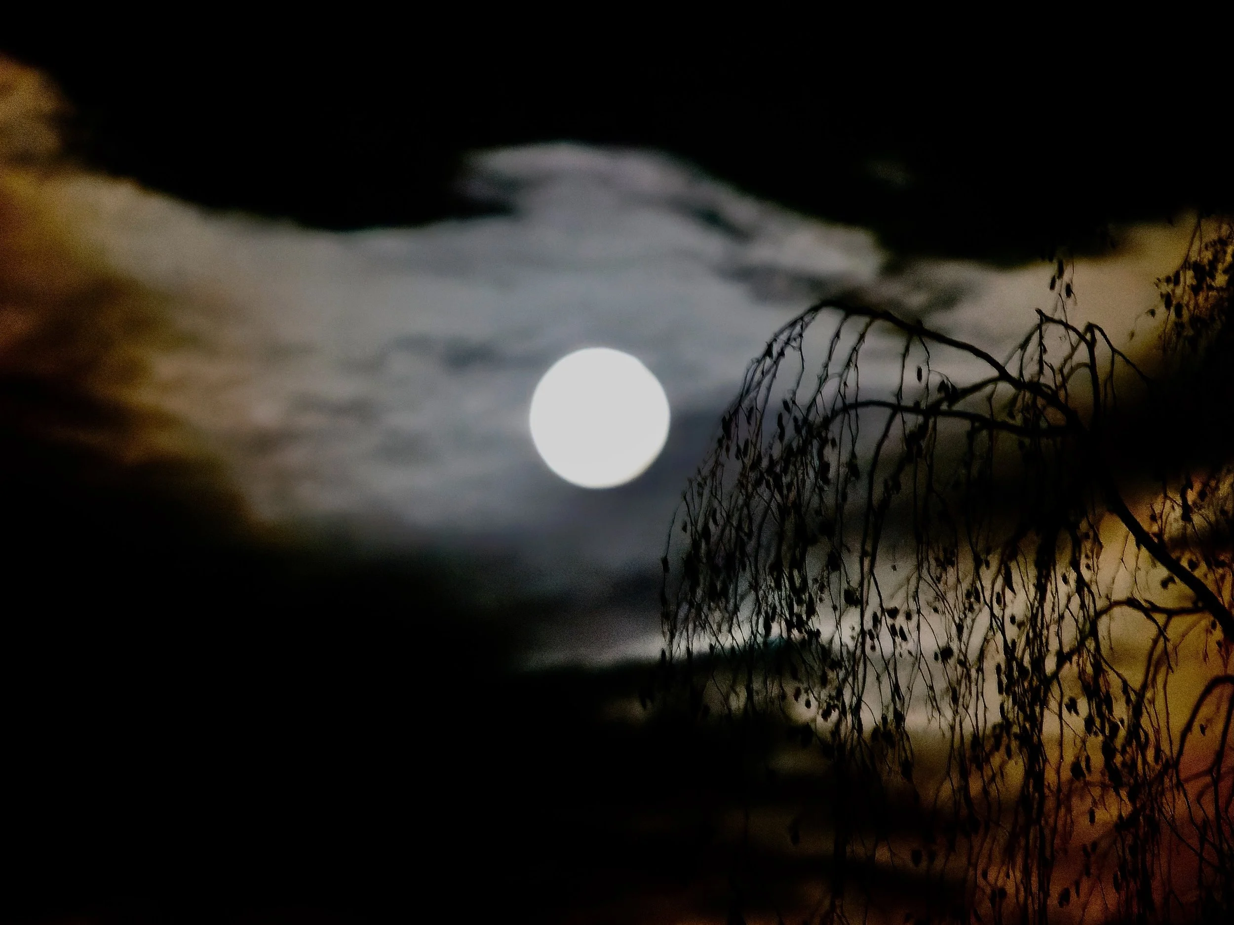 Une pleine lune brillante dans un ciel nuageux, avec des branches d'arbre sans feuilles en premier plan et une lumière orangée à droite.