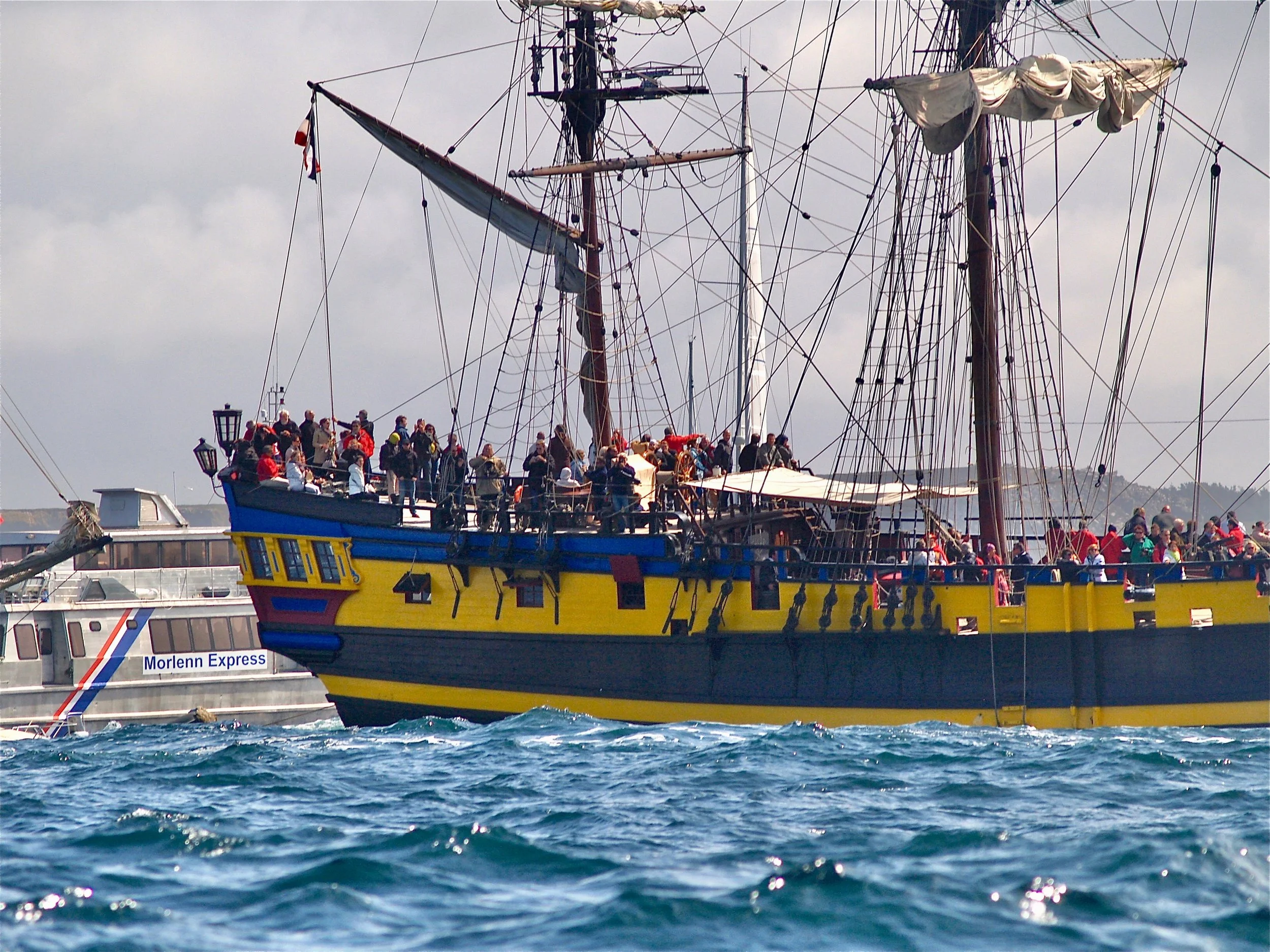 Un grand navire en bois avec de nombreuses cordages et voiles est amarré dans un port. Il y a beaucoup de personnes sur le pont. En arrière-plan, d'autres bateaux de croisière sont visibles.