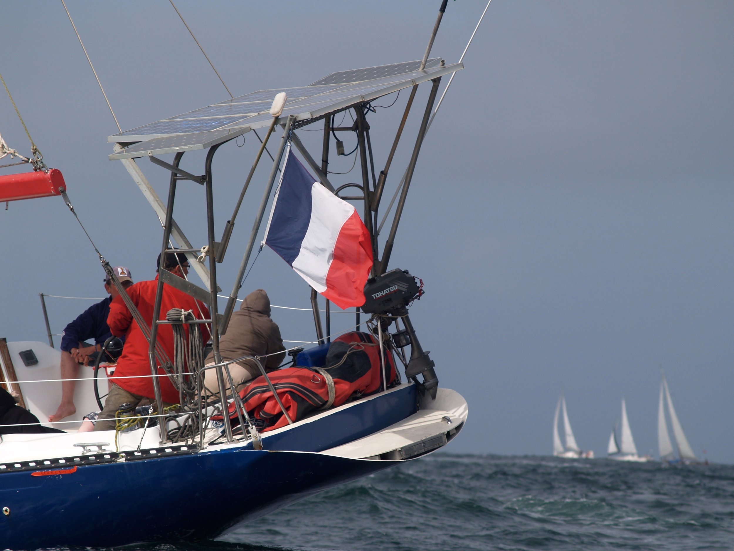 Un voilier avec des gens à bord, dont un porte une veste rouge, et un drapeau français, naviguant en mer sous un ciel nuageux.