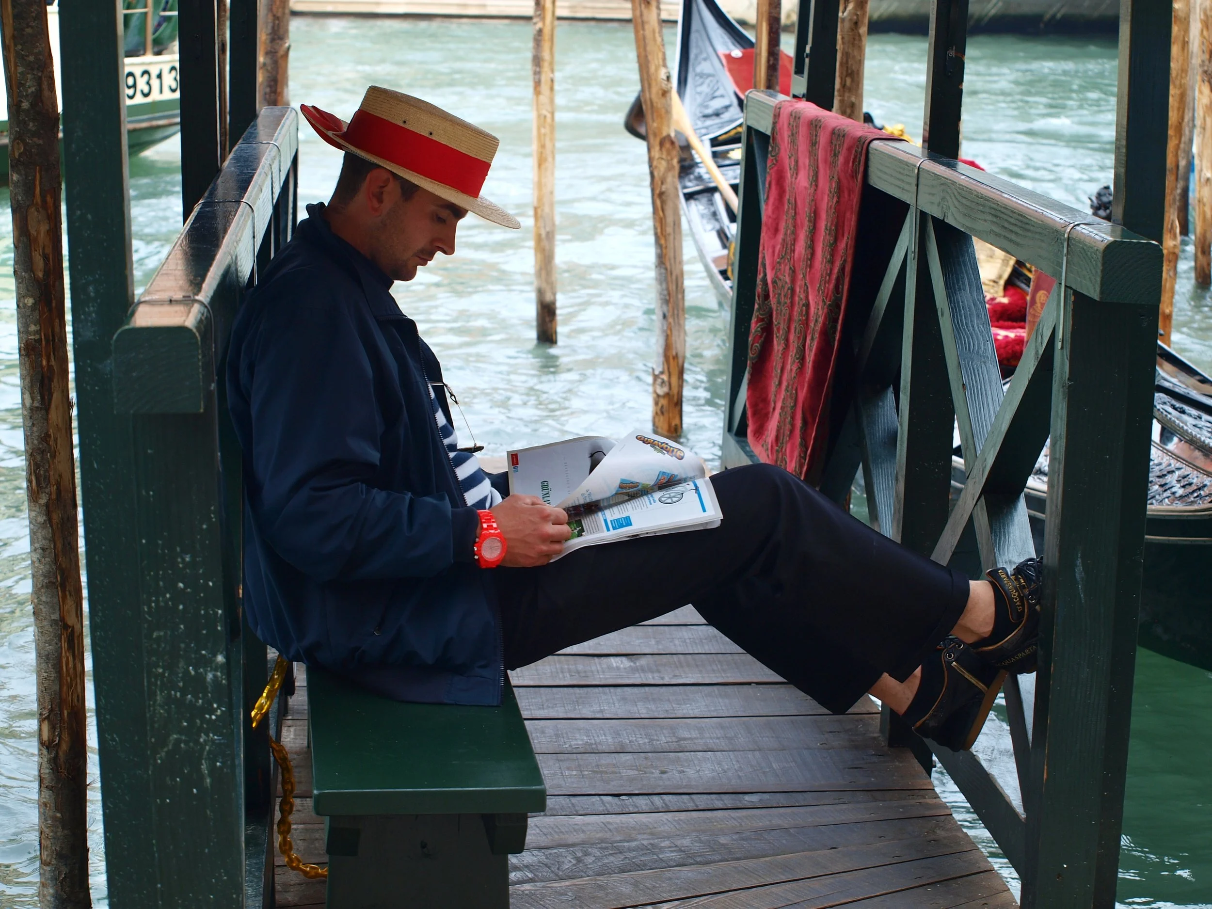 Un homme assis sur un banc en bois sur un quai en bois, portant un chapeau de paille avec un bande rouge, lisant un magazine, avec de l'eau en arrière-plan.