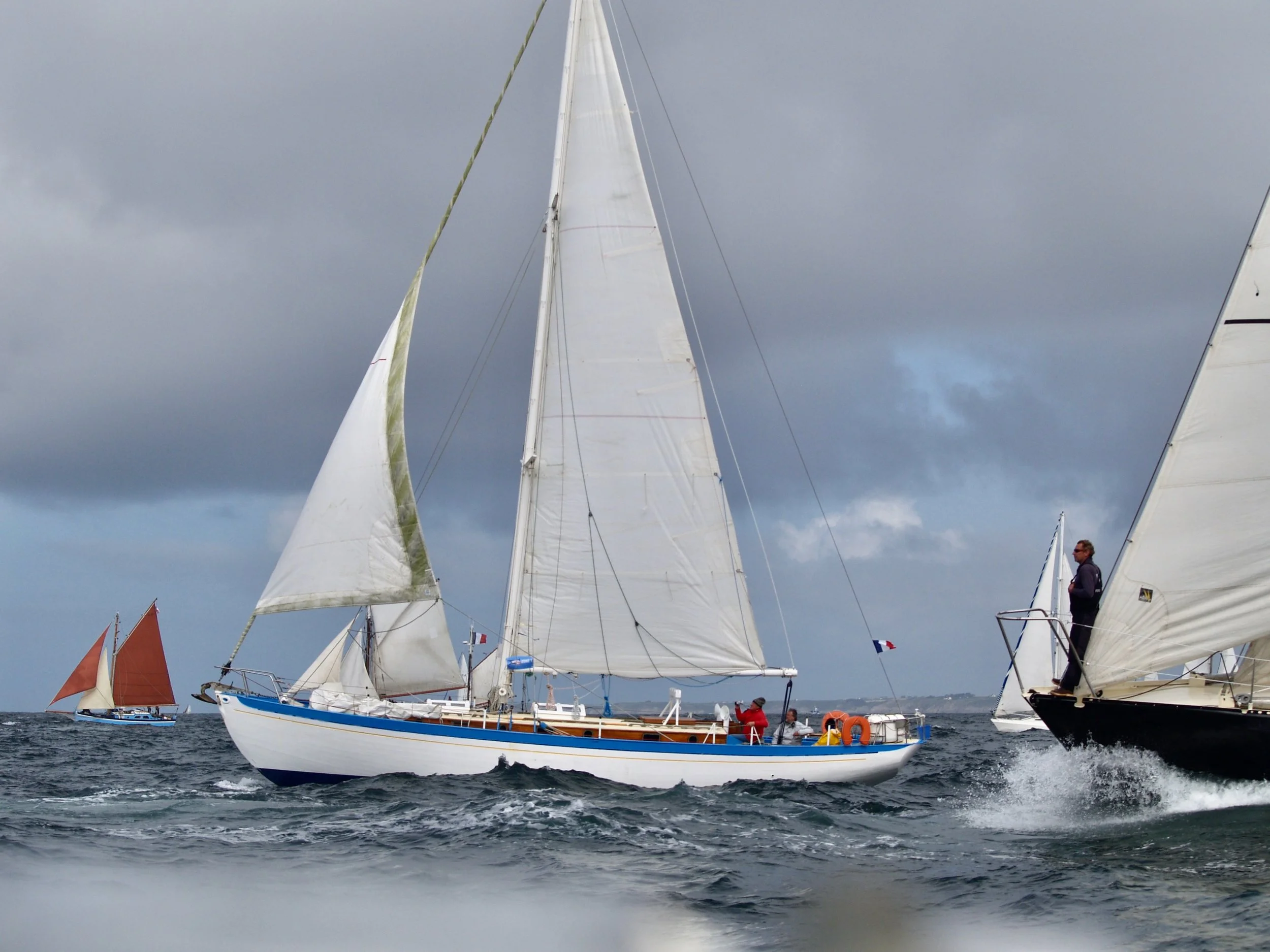 Voilier blanc avec voile large naviguant en mer, autres voiliers en arrière-plan, ciel nuageux, deux personnes sur le pont du voilier.