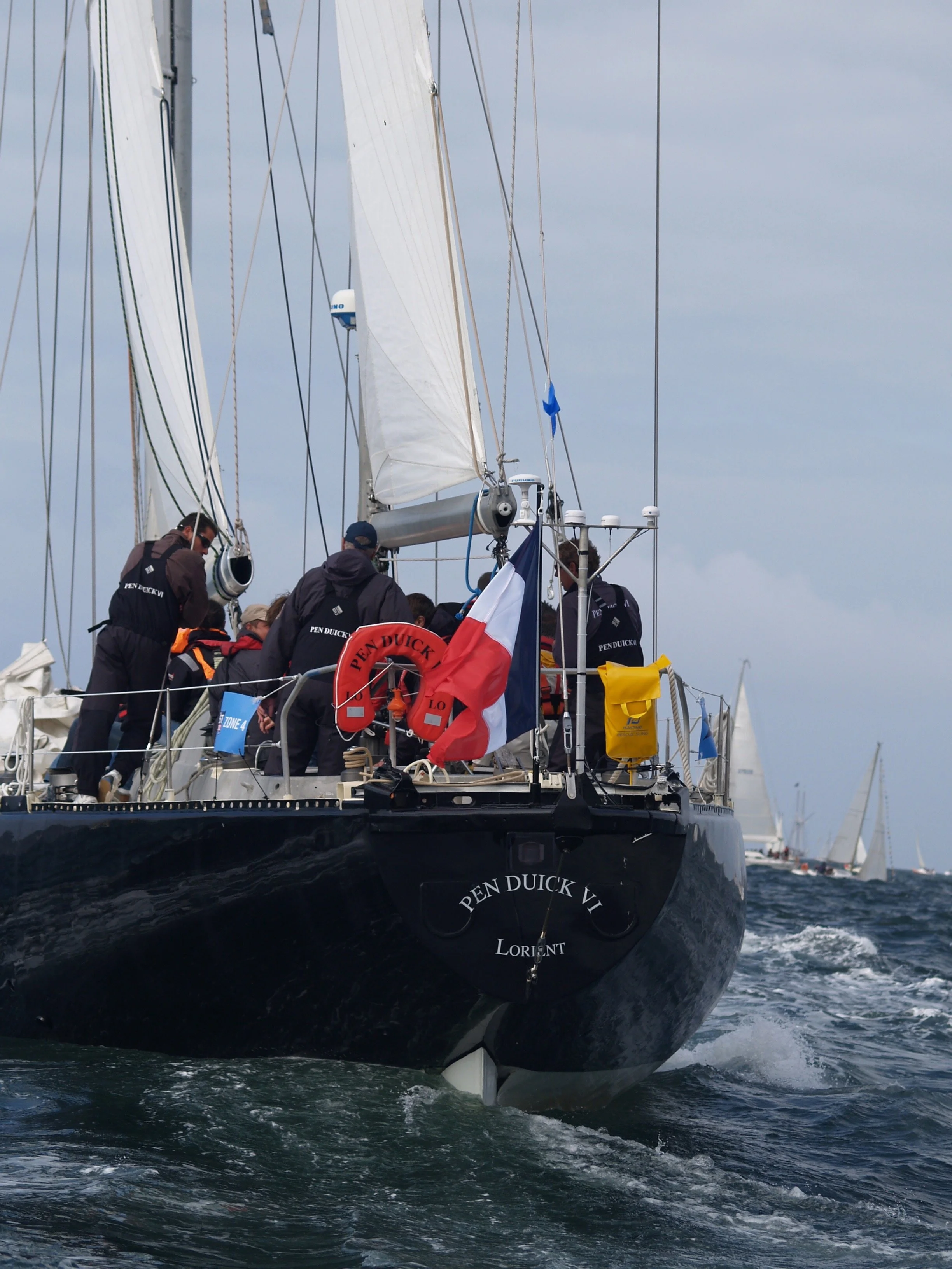 Un voilier participant à une course, avec plusieurs membres d'équipage à bord, en mer avec un ciel nuageux.