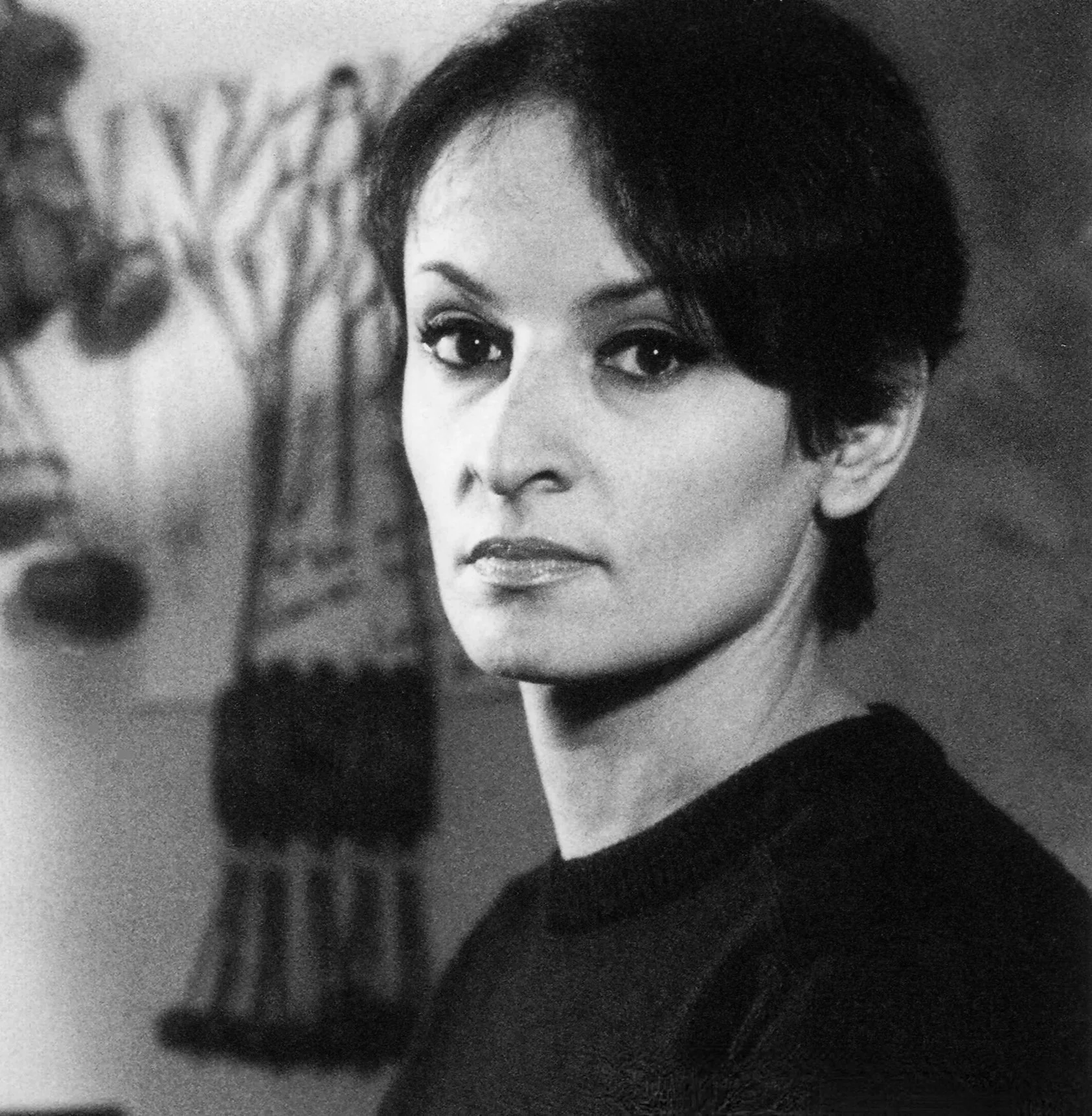 Portrait en noir et blanc d'une femme avec des cheveux courts et un regard direct, portant un vêtement sombre.
